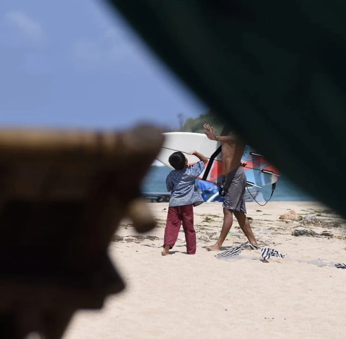 Children at the beach, one helping the other hold a surfboard, seen through a partially obstructed view.