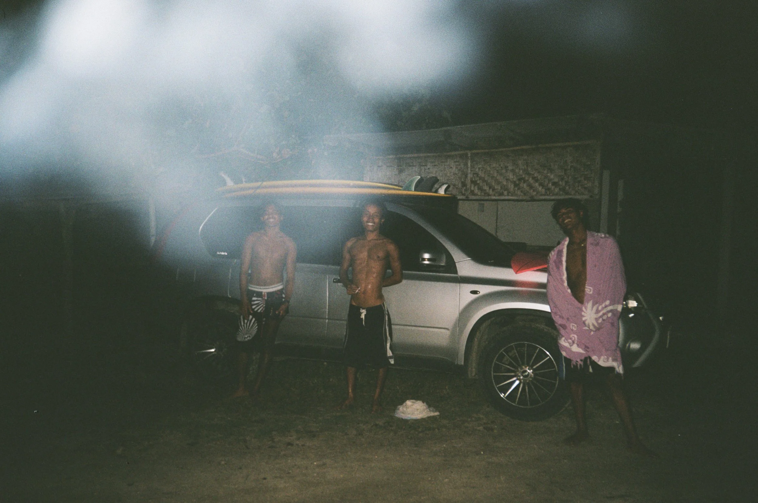 Three young men standing in front of a silver SUV with surfboards on top, night setting, smiling at camera.