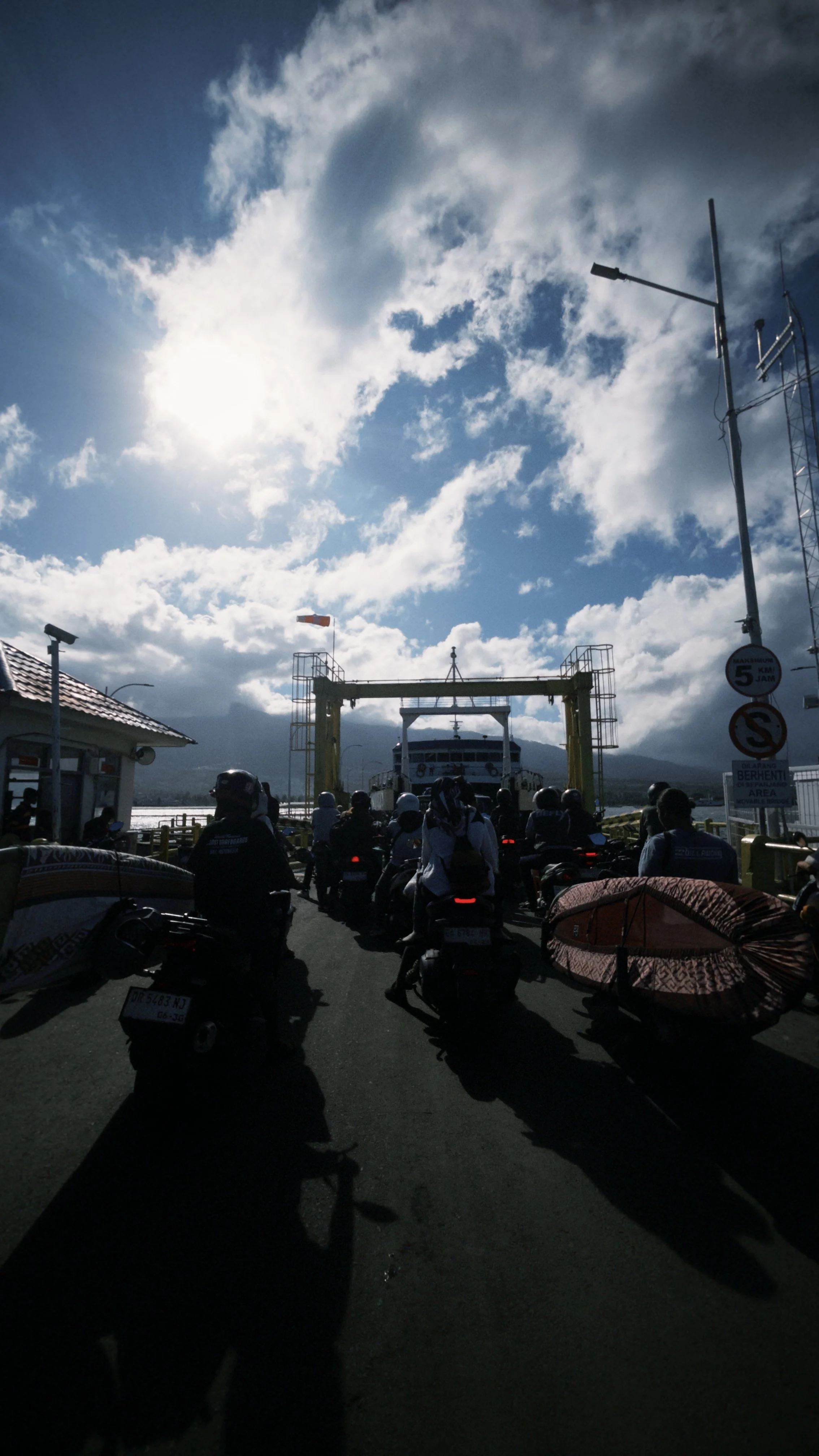 Many people on scooters waiting to board a ferry at a dock, with a cloudy sky and mountains in the background.