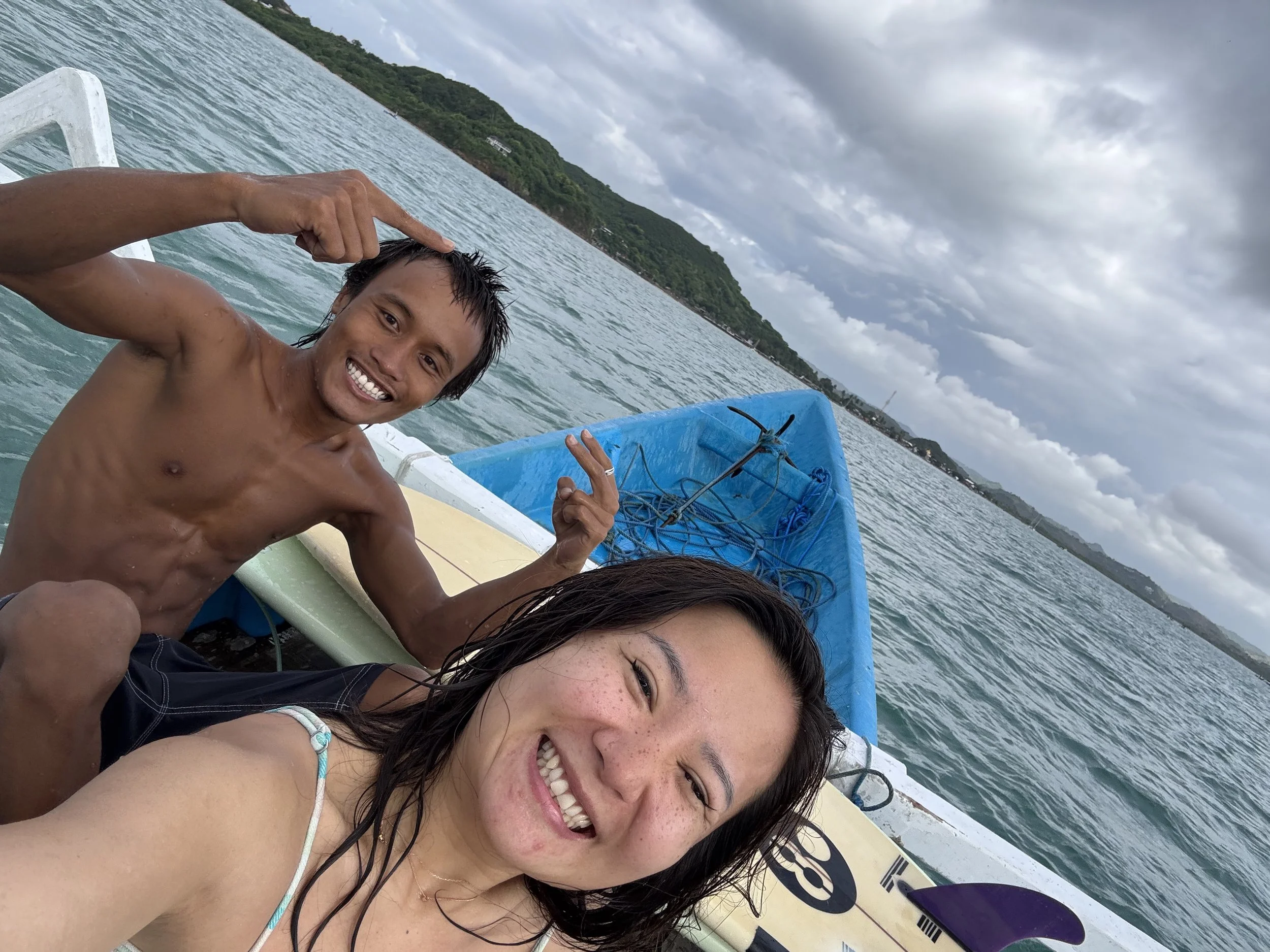 Three people smiling and enjoying a day on a boat, with one person making a peace sign and another shading their eyes, near a hilly coastline under cloudy skies.