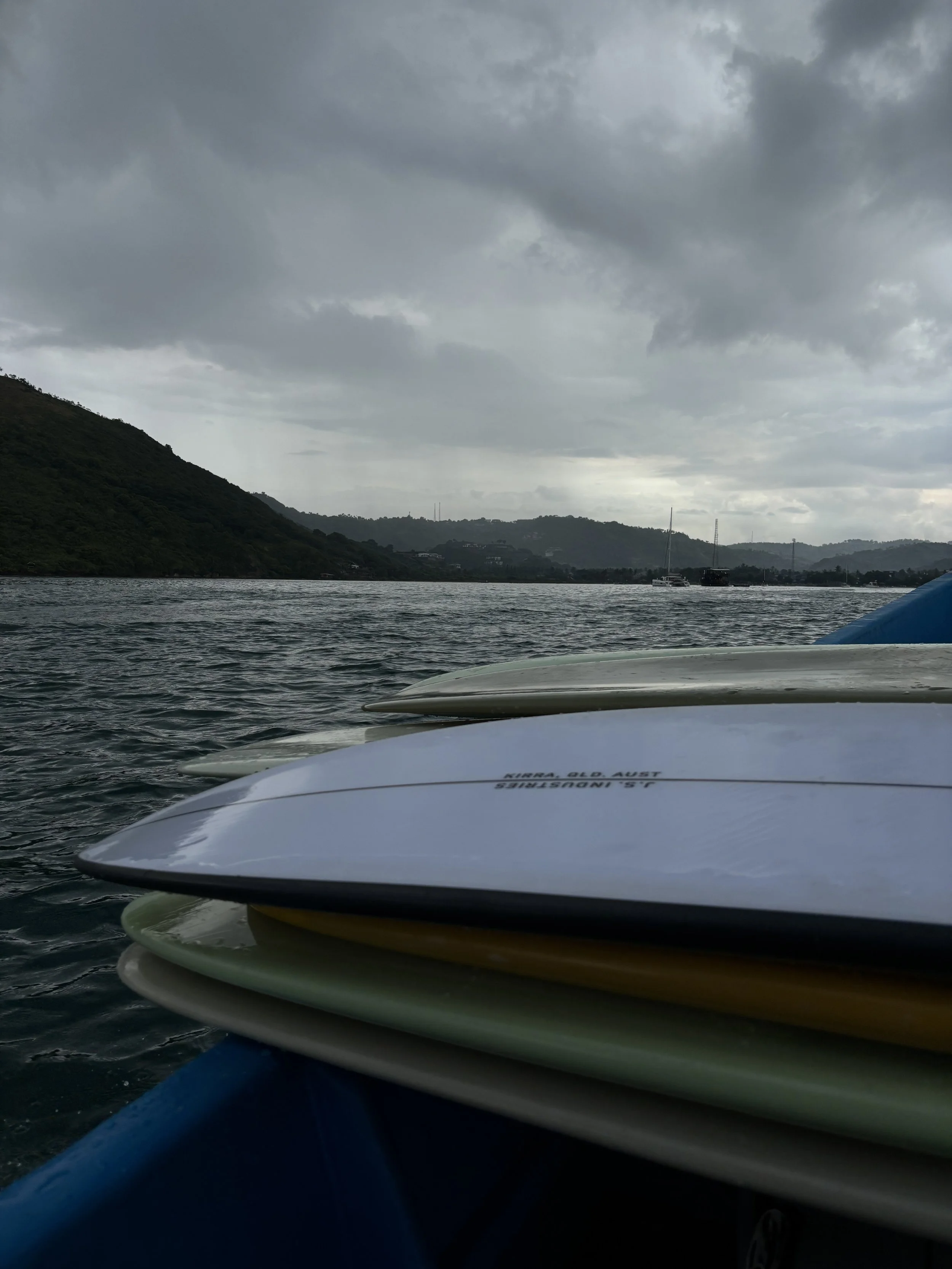 Close-up of several stacked surfboards on a boat, with a body of water and a cloudy sky in the background.