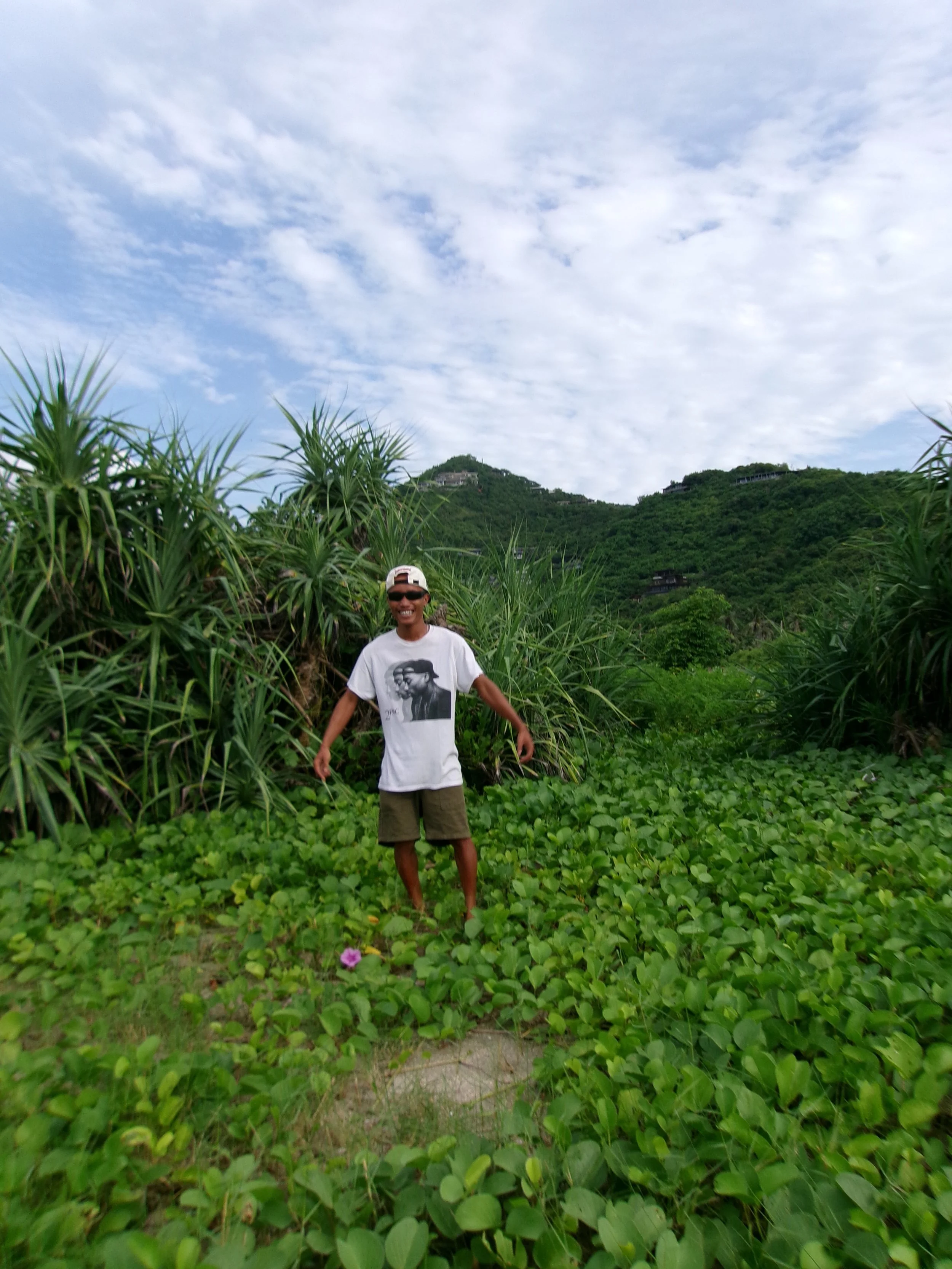 A person wearing sunglasses, a white t-shirt with a black and white portrait, and a white cap, standing in a lush green area with dense plants, with a mountain or hill covered in greenery in the background under a cloudy sky.