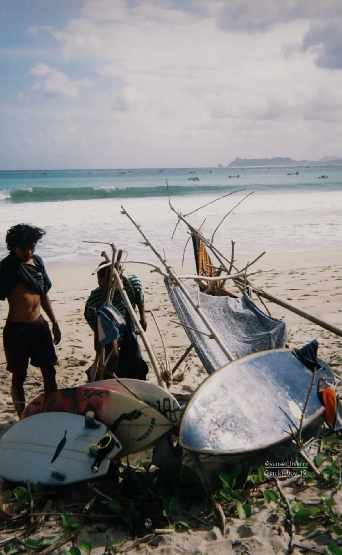 Two children and a man standing near broken surfboards and a makeshift shelter made from sticks on a sandy beach with the ocean and distant land in the background.