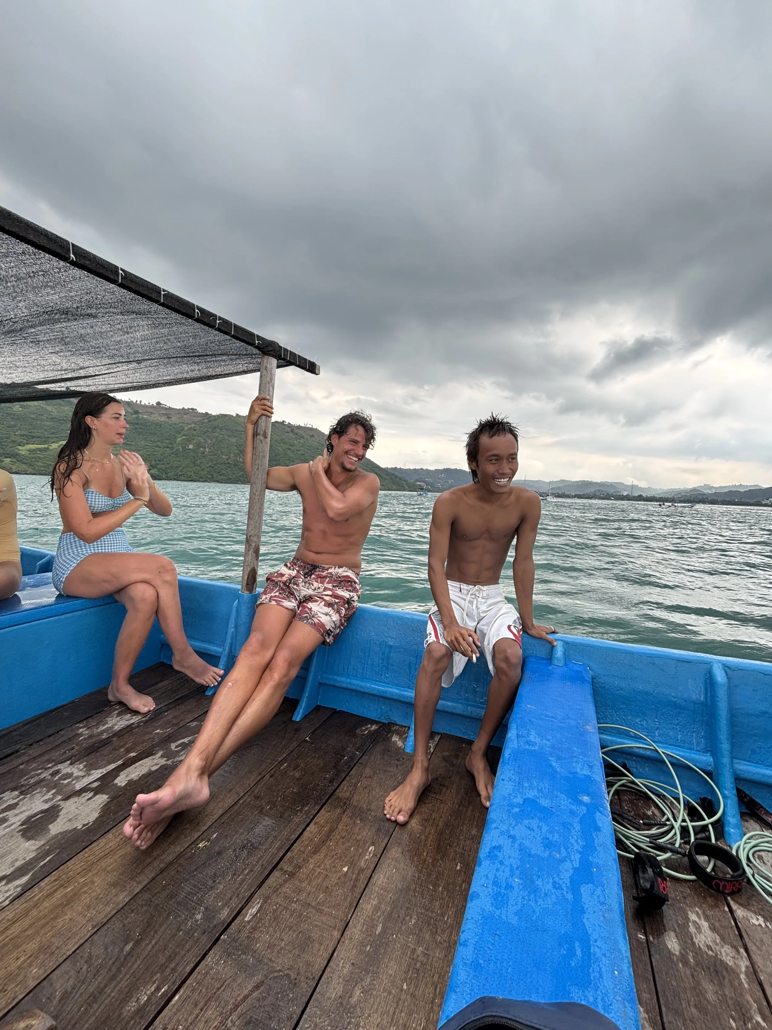 People sitting and standing on a boat with a wooden deck, some smiling and one woman in a blue checkered outfit, with a cloudy sky and hills in the background.