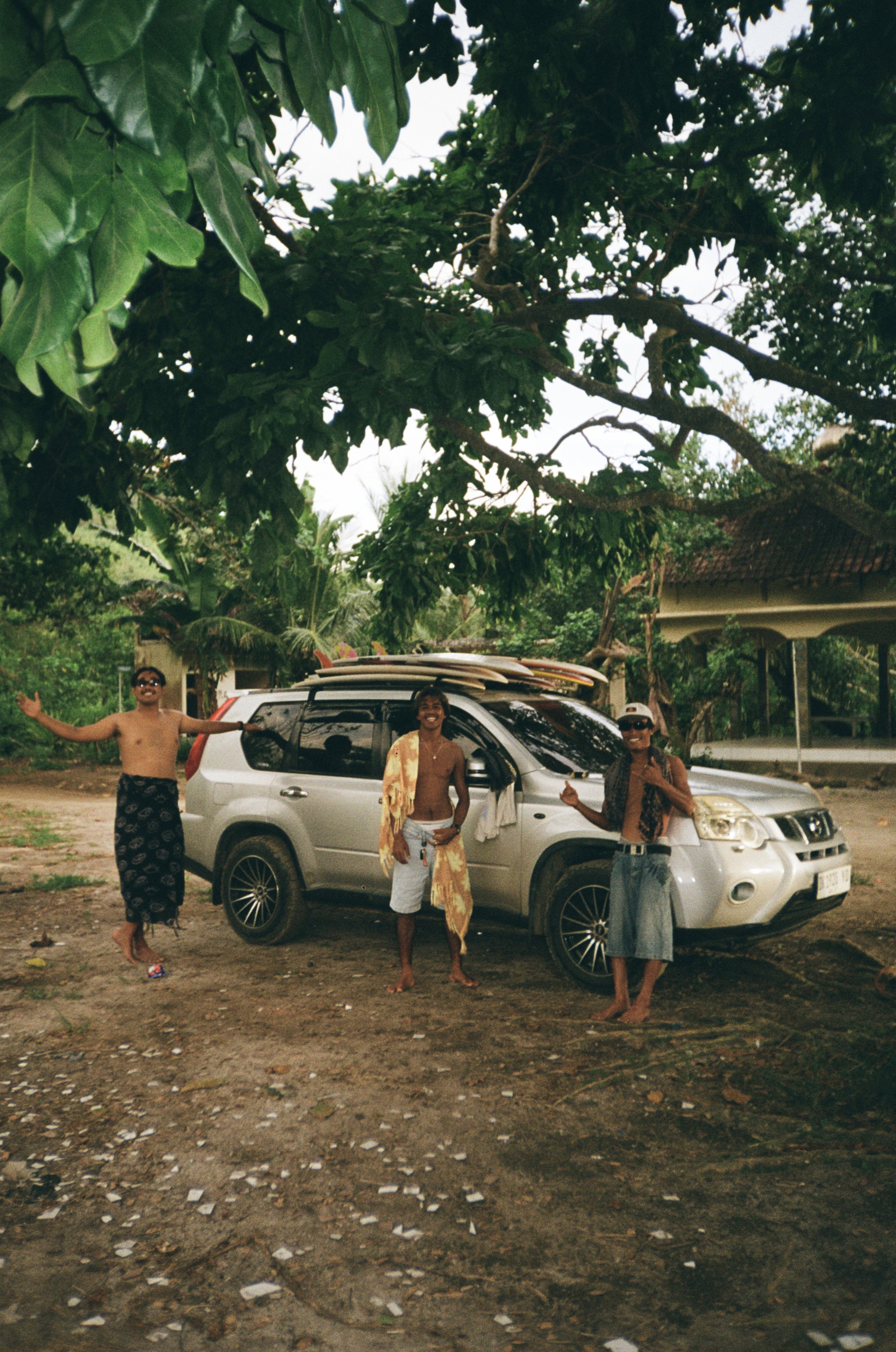 Three people are standing in front of a silver SUV under a large tree with broad green leaves. Two of them are shirtless and wearing sarongs, while the third person is wearing a sleeveless top, sunglasses, and jeans. They appear to be enjoying themselves outdoors on a rural or semi-rural property with a building visible in the background.