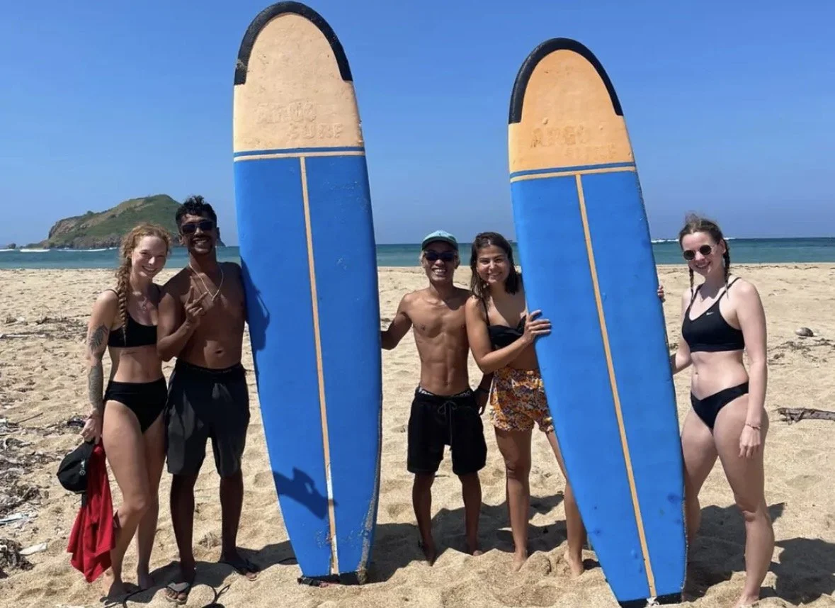 Group of five friends on the beach with surfboards, smiling and posing for the photo.