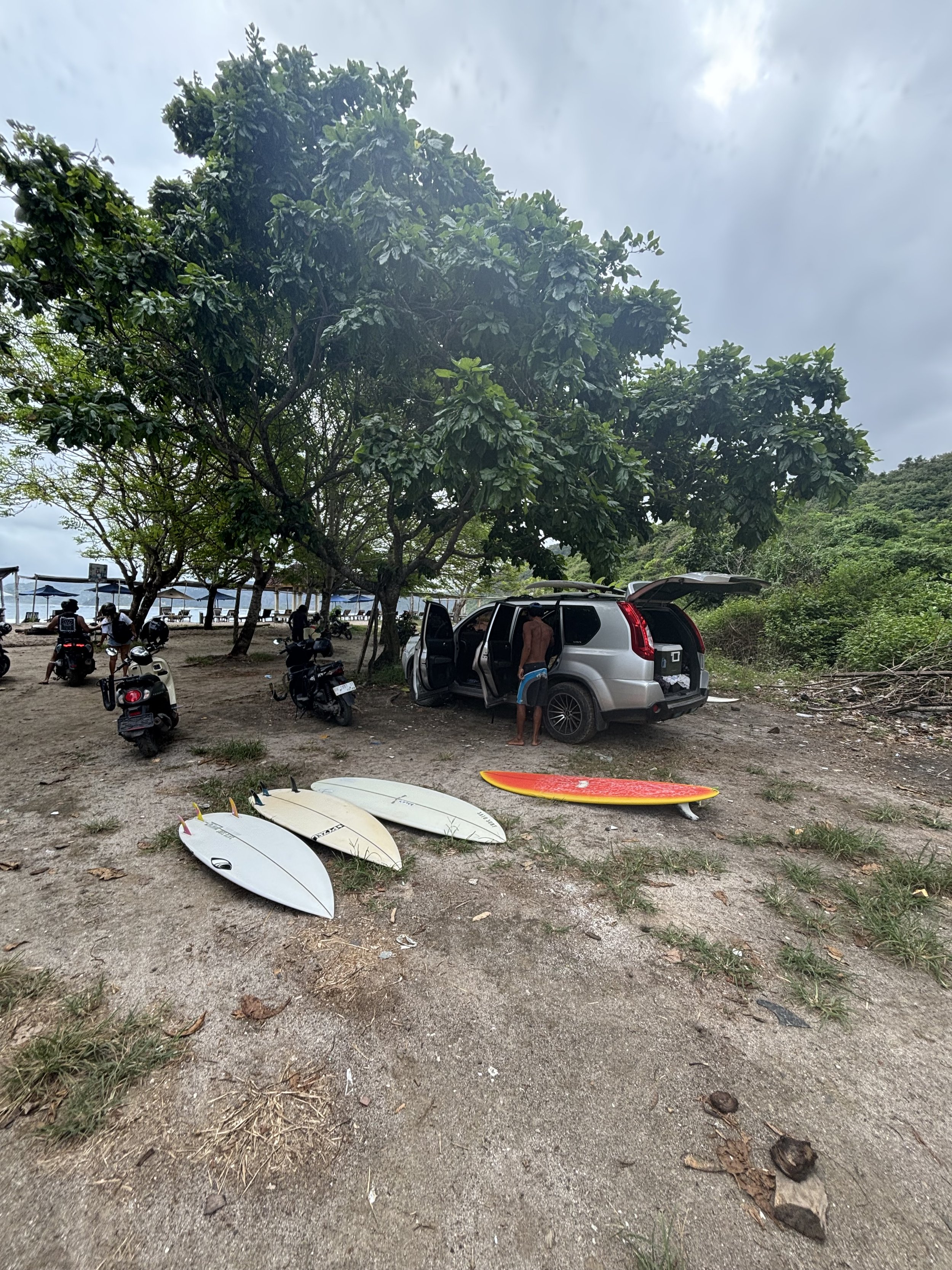 Beach scene with several surfboards on the ground, large green trees, a silver SUV with open trunk, and people near the shoreline, cloudy sky overhead.