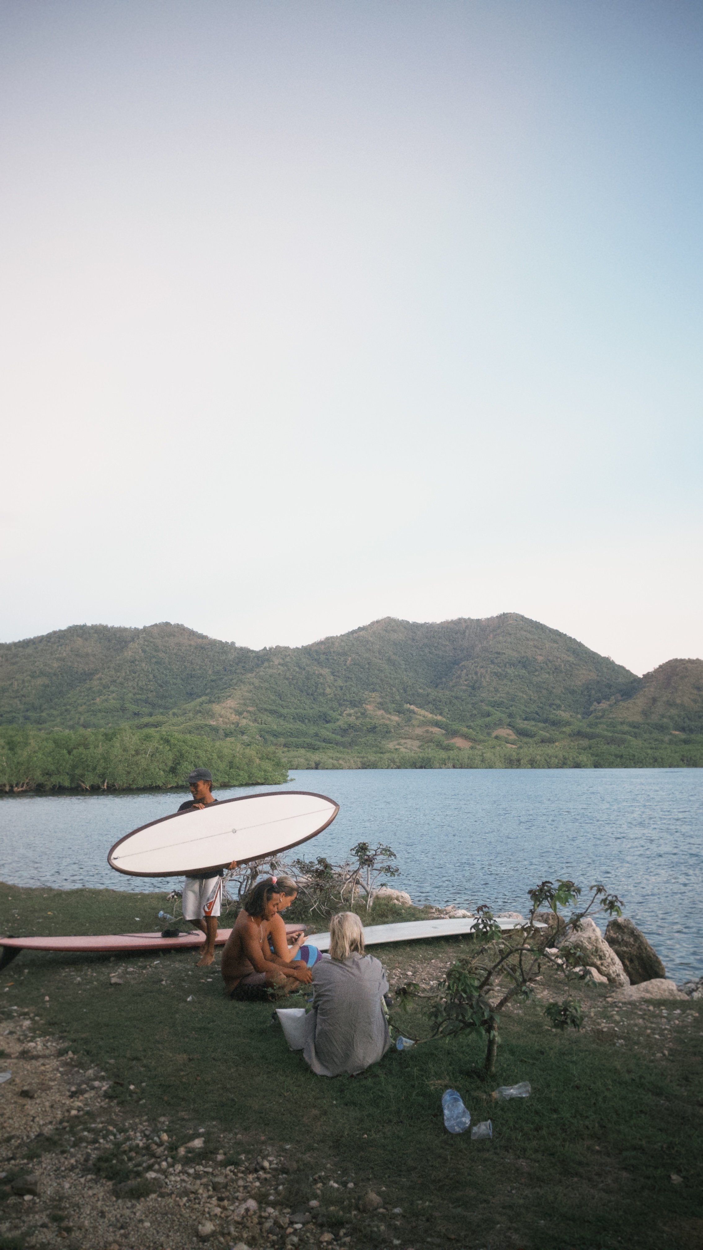 People relaxing by a lake with mountains in the background, some holding surfboards, and trees and rocks in the foreground.