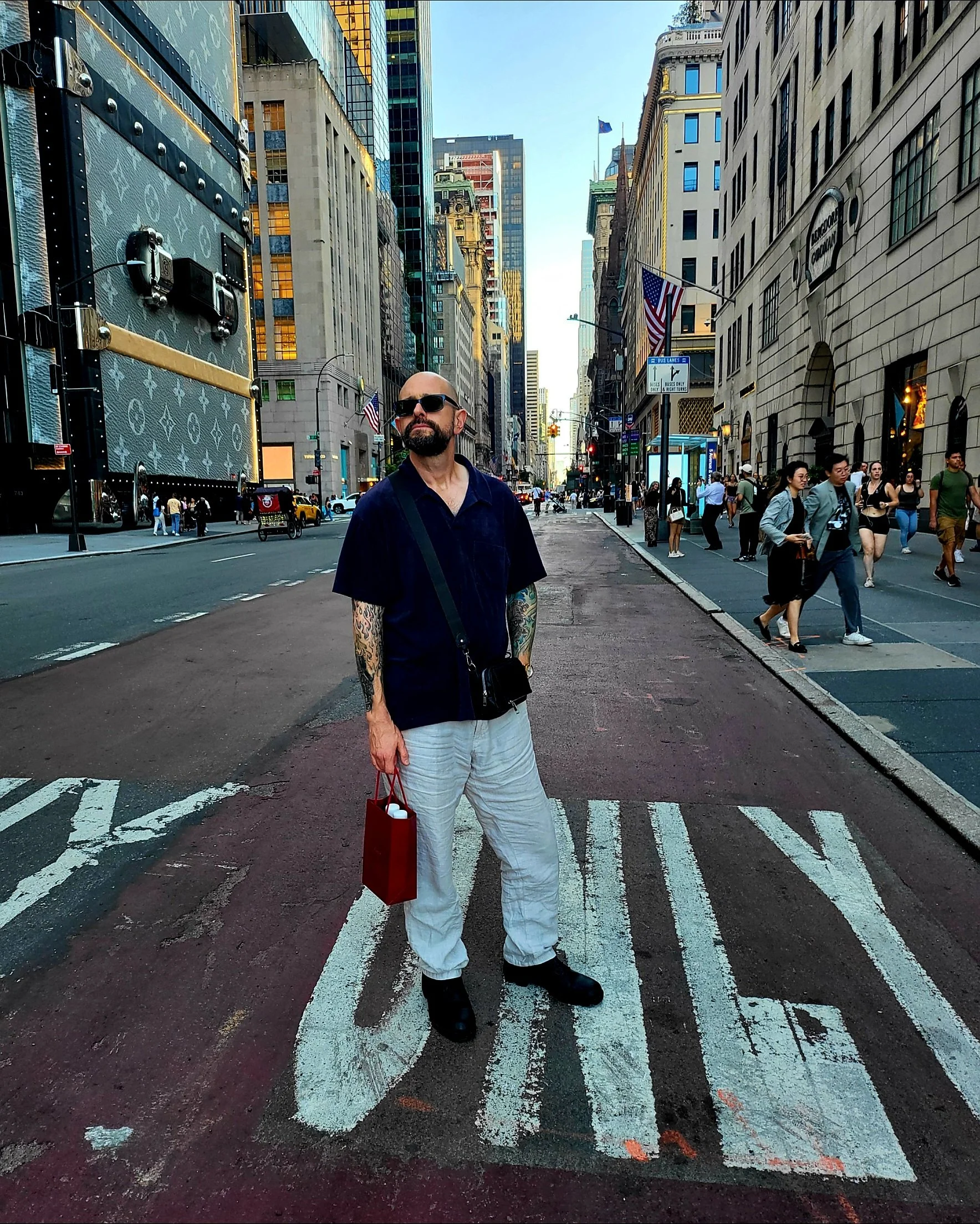 A man wearing sunglasses, a navy blue shirt, and white pants stands on a city street with tall buildings and people walking by. He holds a red shopping bag and looks to his left.