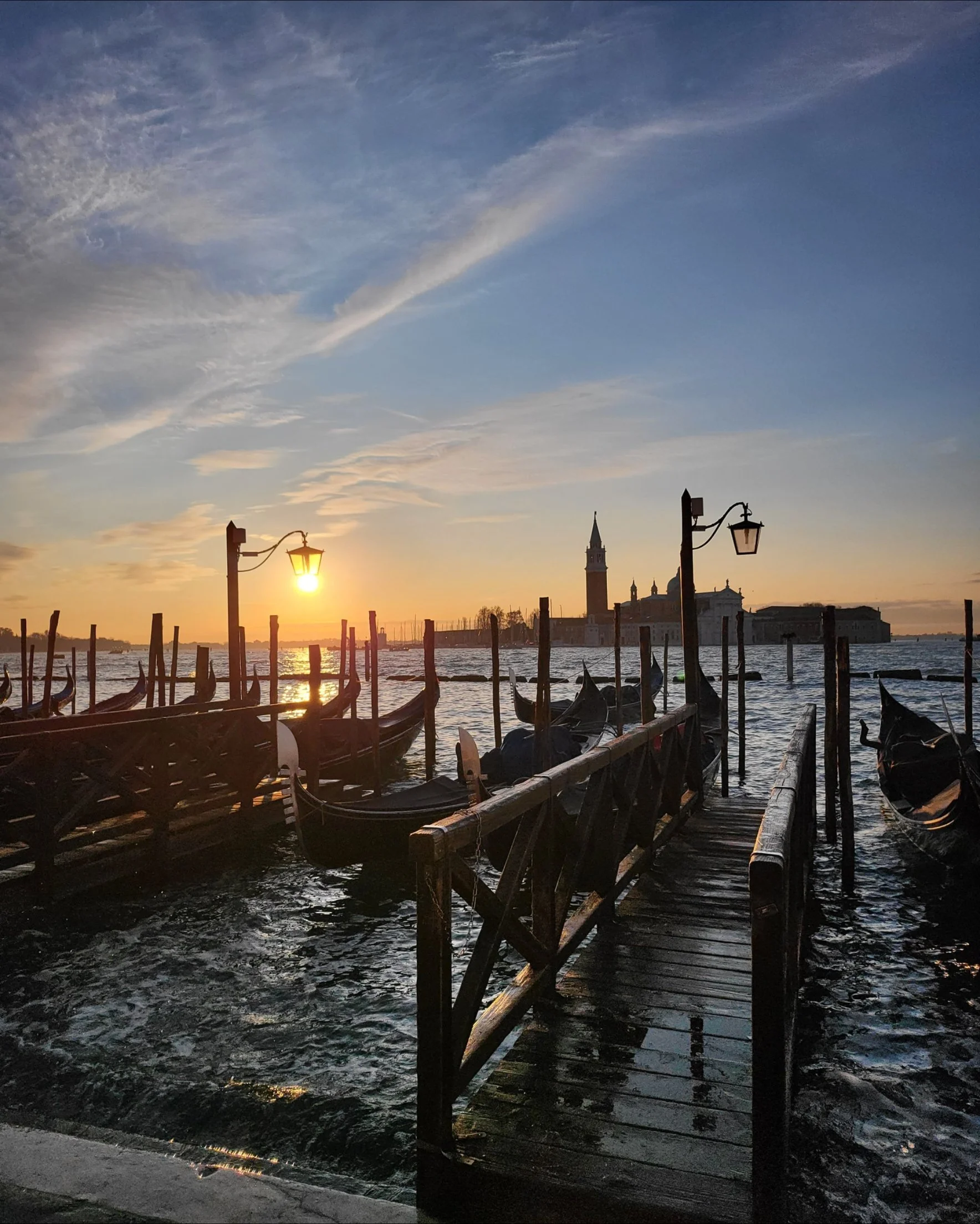 Sunset over a waterfront with gondolas docked along a wooden pier, old-fashioned street lamps, and silhouettes of historic buildings and a church steeple in the distance.