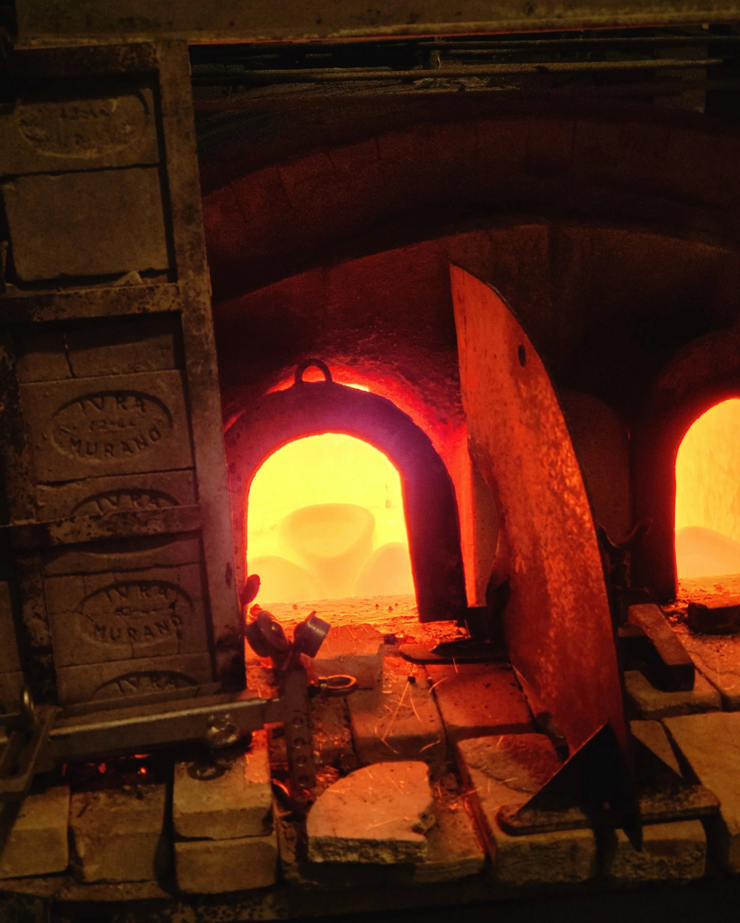Close-up of a wood-fired oven with glowing red-orange flames visible through the opening, surrounded by brickwork and metal frames.