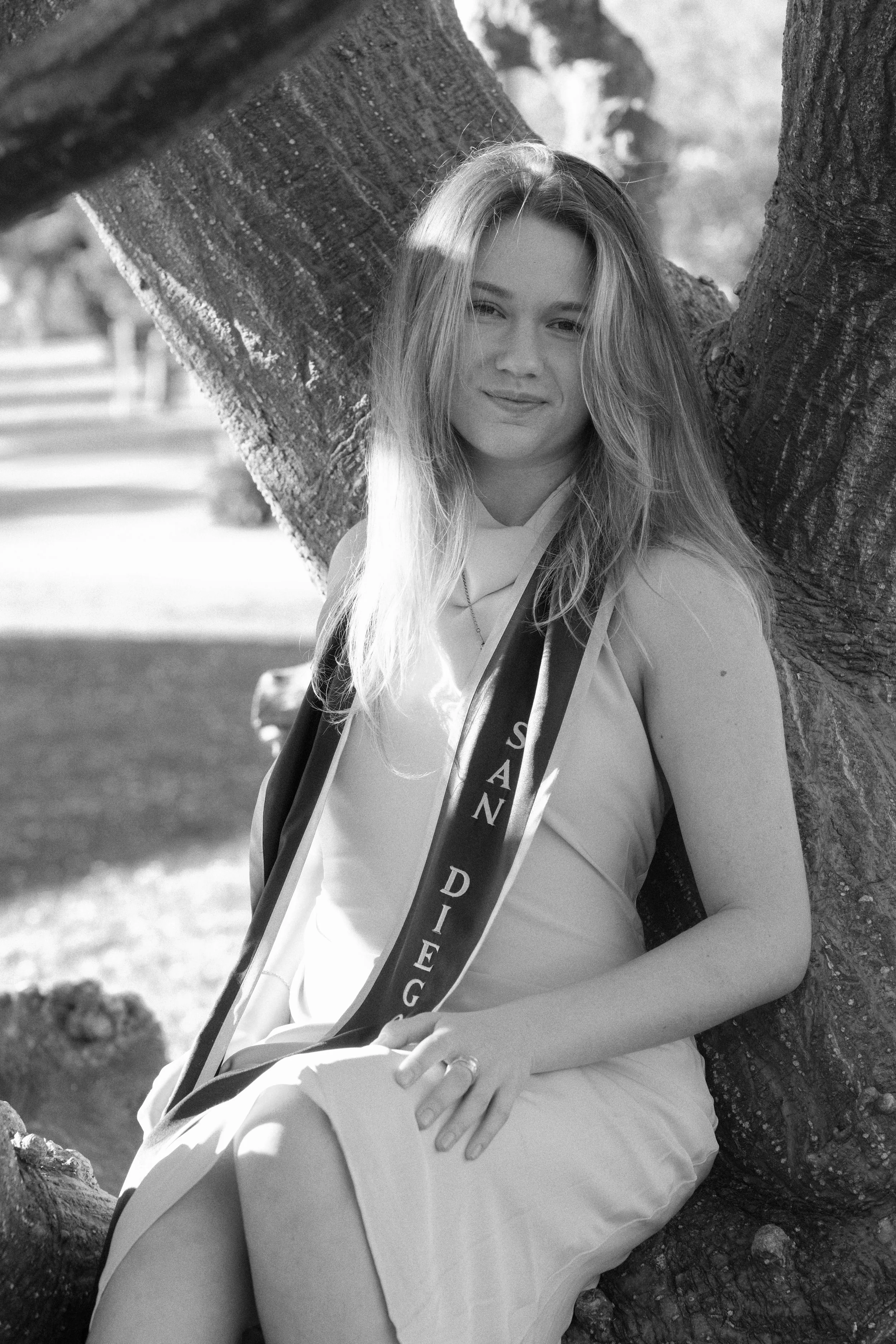A young woman with long, wavy hair sitting under a large tree, smiling gently at the camera. She is wearing a sleeveless dress and a sash that reads 'SANDIEGO,' likely in celebration of a graduation or similar event.