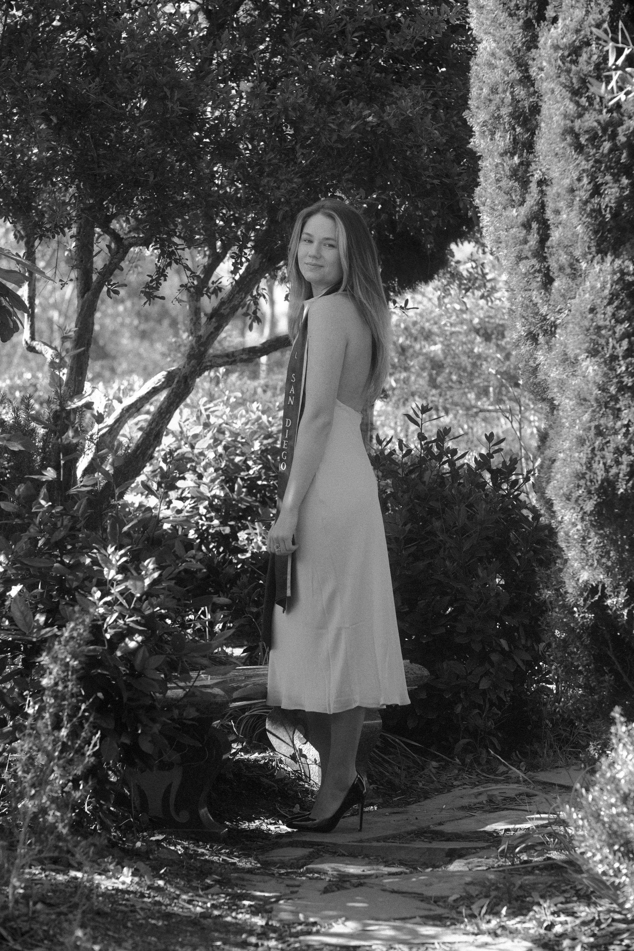 A woman wearing a sash and high heels standing outdoors among trees and bushes in a garden or park. She is looking at the camera with a slight smile.