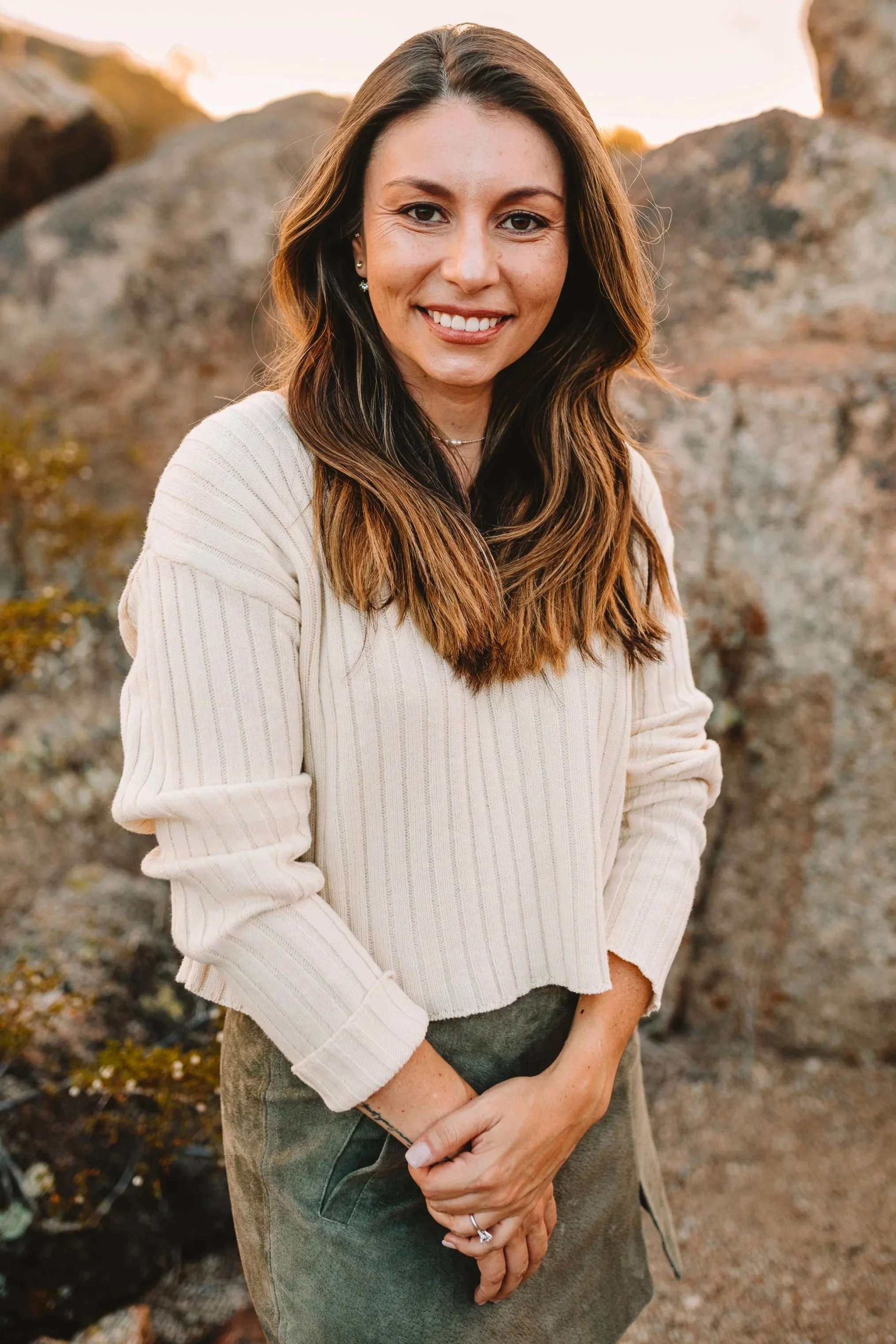 A woman with long brown hair smiling outdoors, wearing a cream sweater and green pants, in front of rocky terrain at sunset.