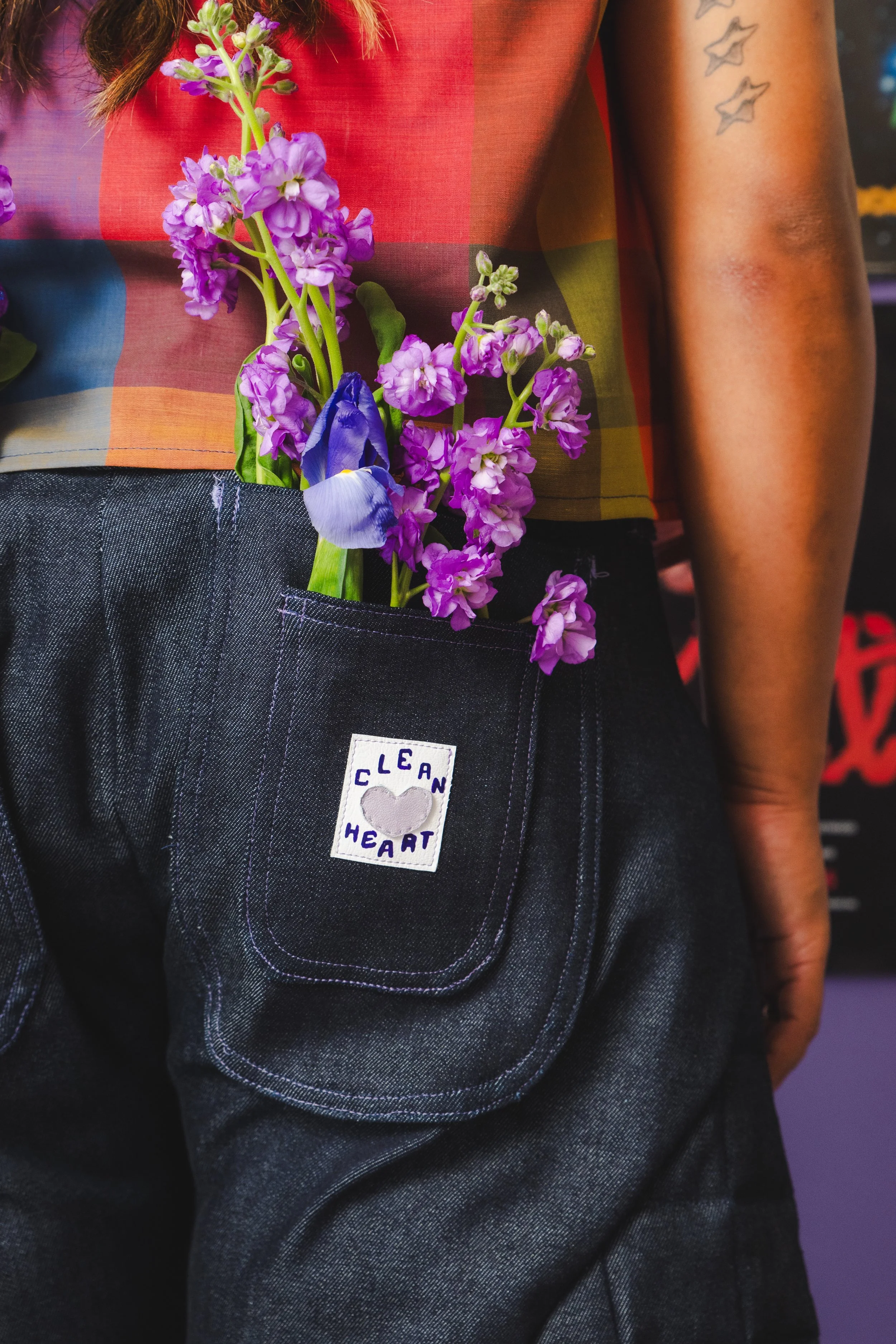 A person wearing a red, yellow, and green checkered shirt and dark denim overalls with a patch that says 'Clean Heart' and a small heart symbol. They have purple flowers, possibly irises and stock flowers, tucked into their pocket and held behind the shirt. The person's right arm, which has small star tattoos, is visible.
