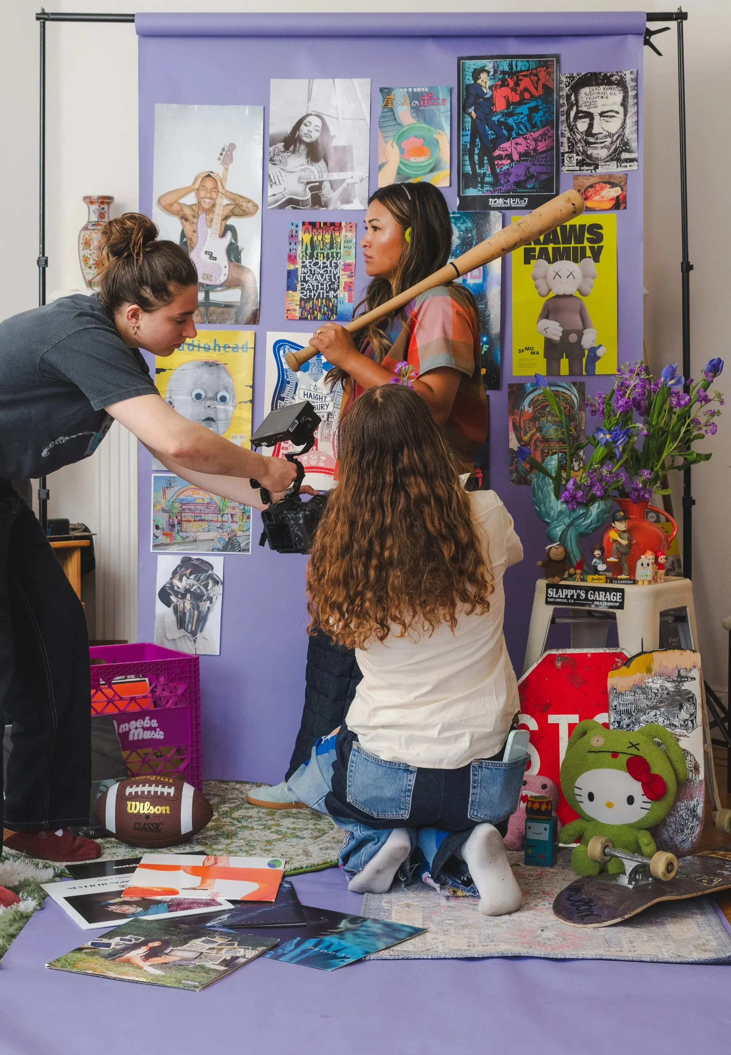 People filming a photo shoot in a room with a lavender backdrop covered in posters and artwork. One woman is crouched, another standing with a baseball bat, a third person is adjusting a camera. The scene includes skateboards, a football, toys, and magazines on the floor.