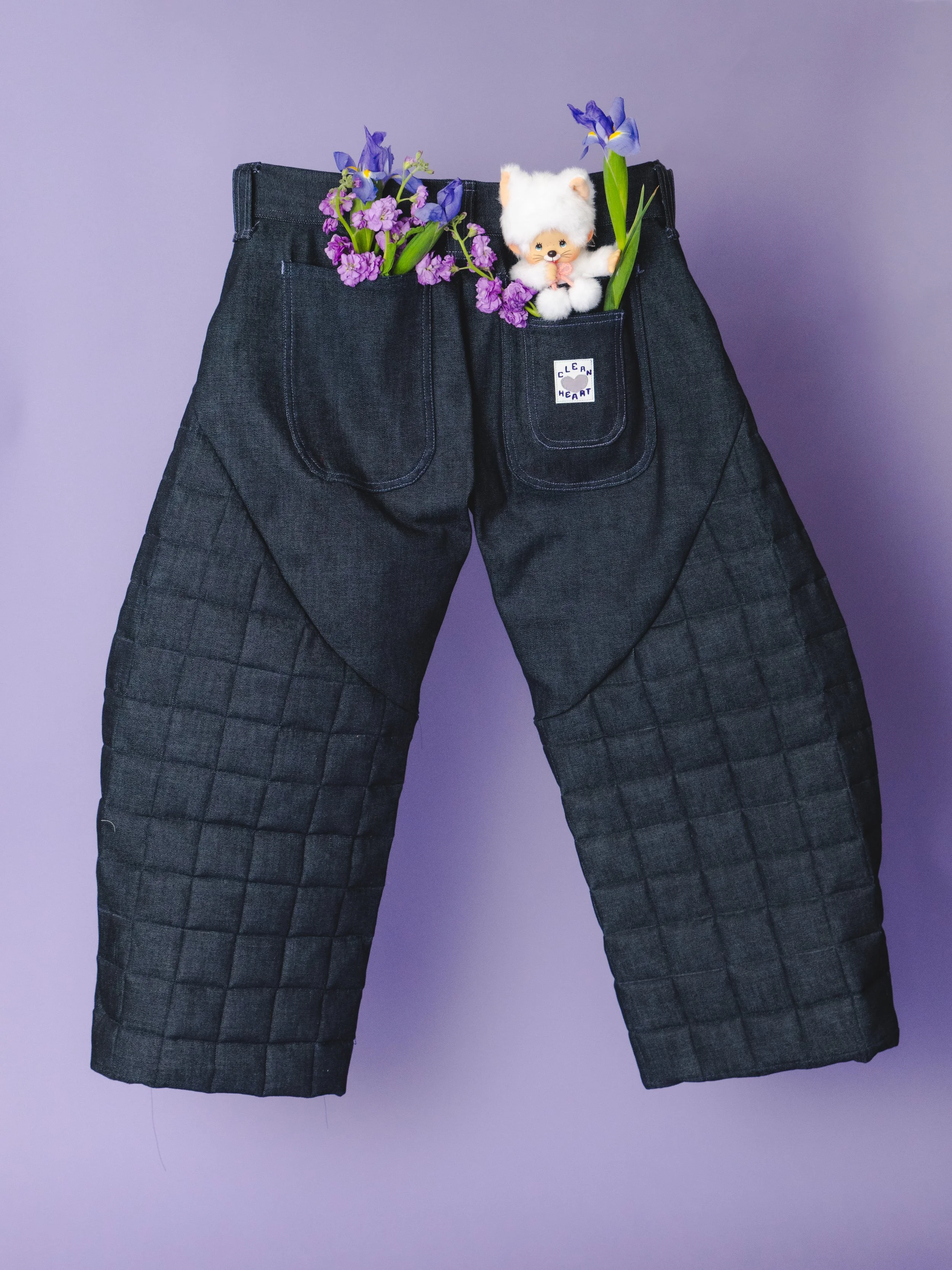 Black quilted pants with front pockets on a purple background, with one pocket containing purple flowers, a small plush toy, and a blue iris flower.