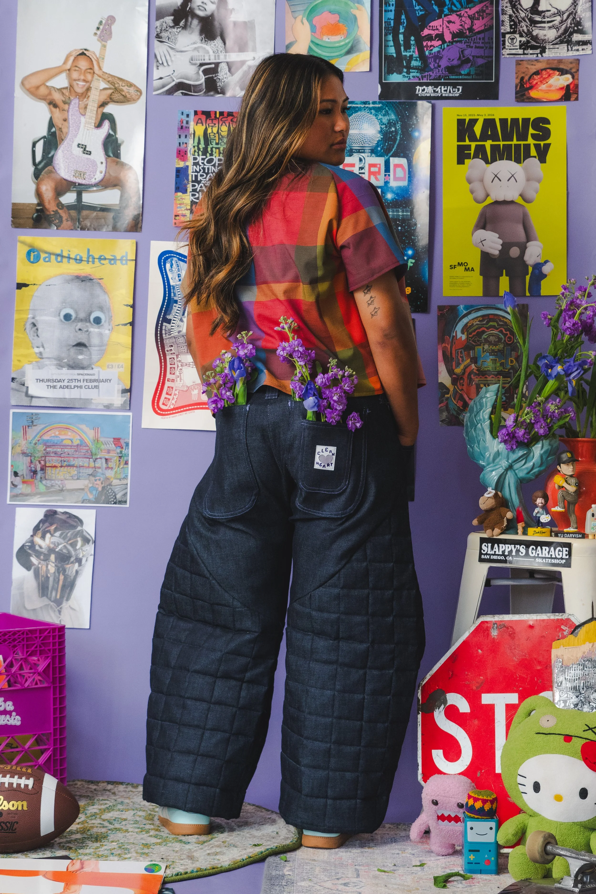 A woman with long wavy hair wearing a colorful checkered shirt and dark wide-legged pants with patches stands in front of a purple wall decorated with posters. She is looking to the right and has tattoos on her left arm. There is a vase with purple f