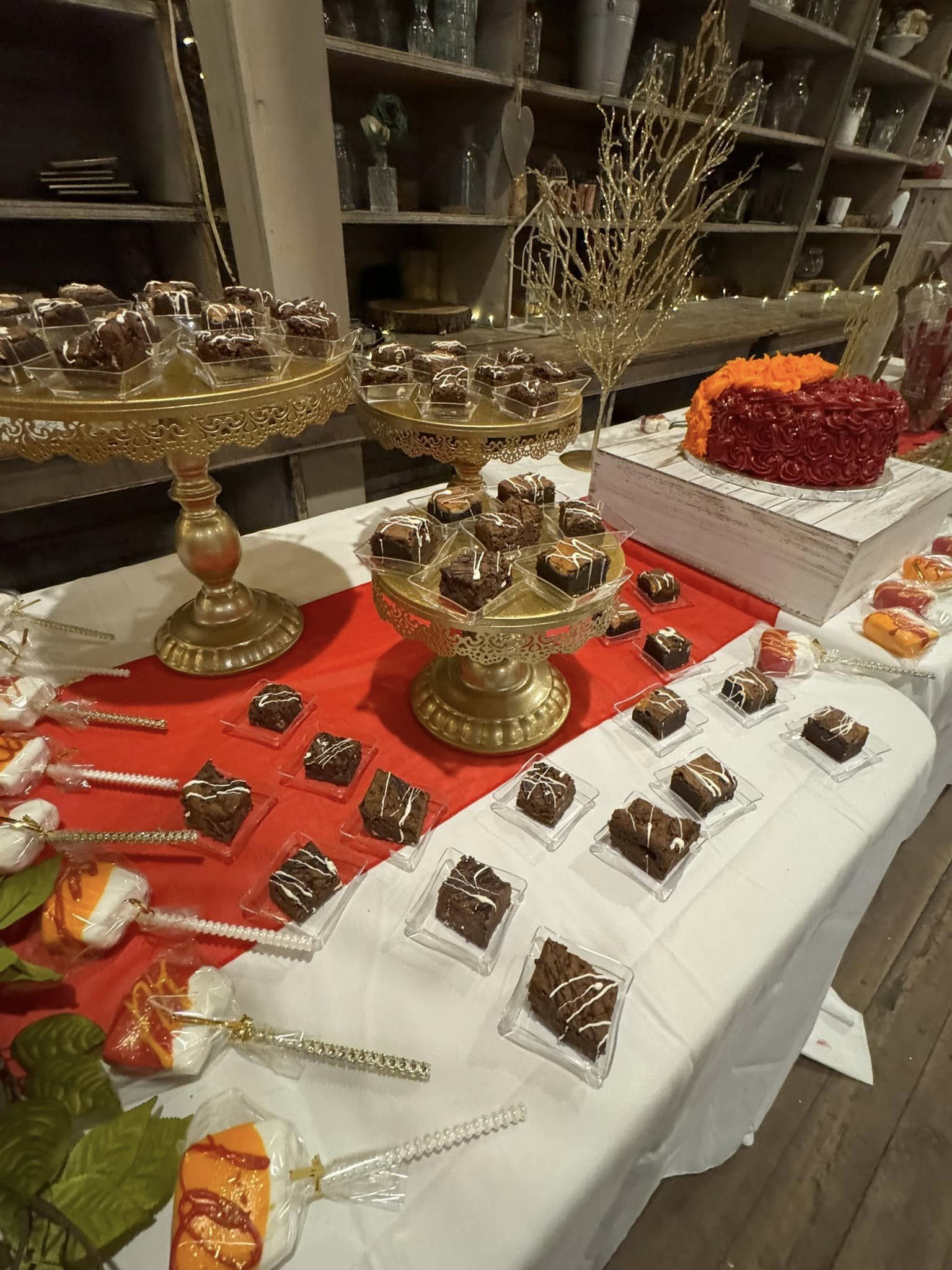 A table decorated with various desserts including brownies and candies, with a large heart-shaped cake topped with orange and red frosting, and gold decorative branches.