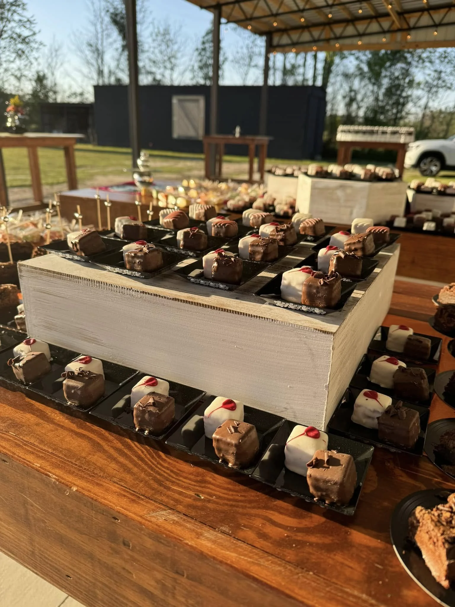 Assorted mini desserts, including chocolate-covered treats with red accents, displayed on black trays at an outdoor event under a pavilion.