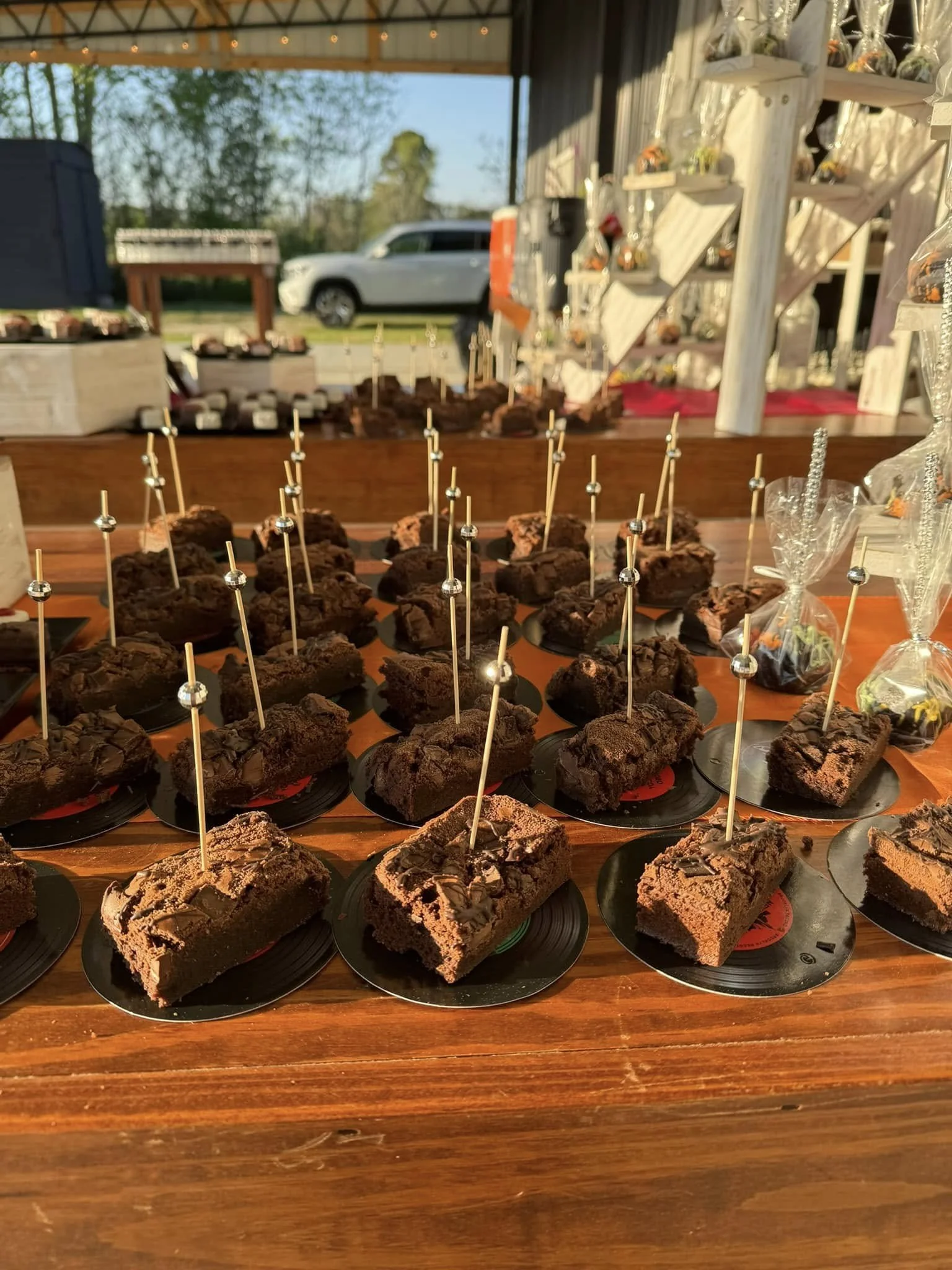 Chocolate brownies with sticks on black plates on a wooden table at an outdoor event, with a display of small treats and decorations in the background.