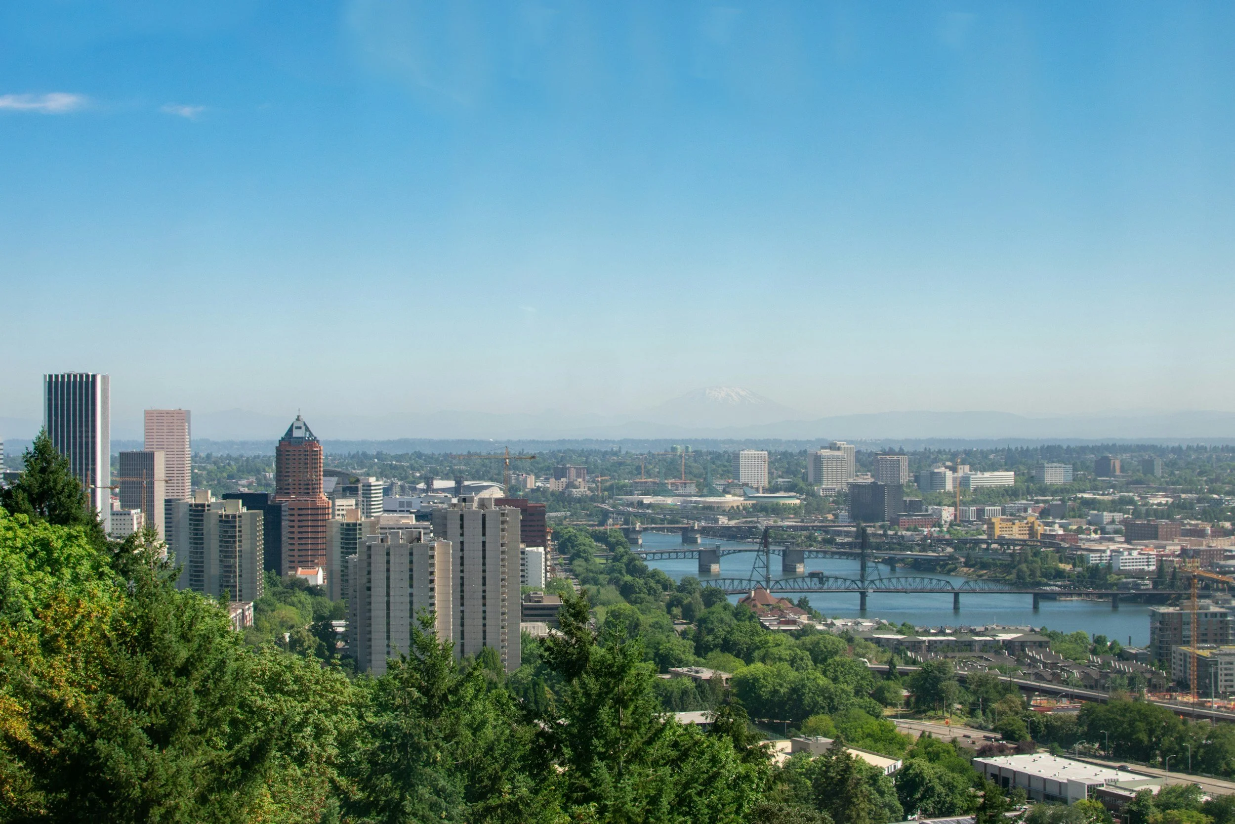 A city skyline with tall buildings, a bridge over a river, and a mountain in the distance under a clear blue sky.