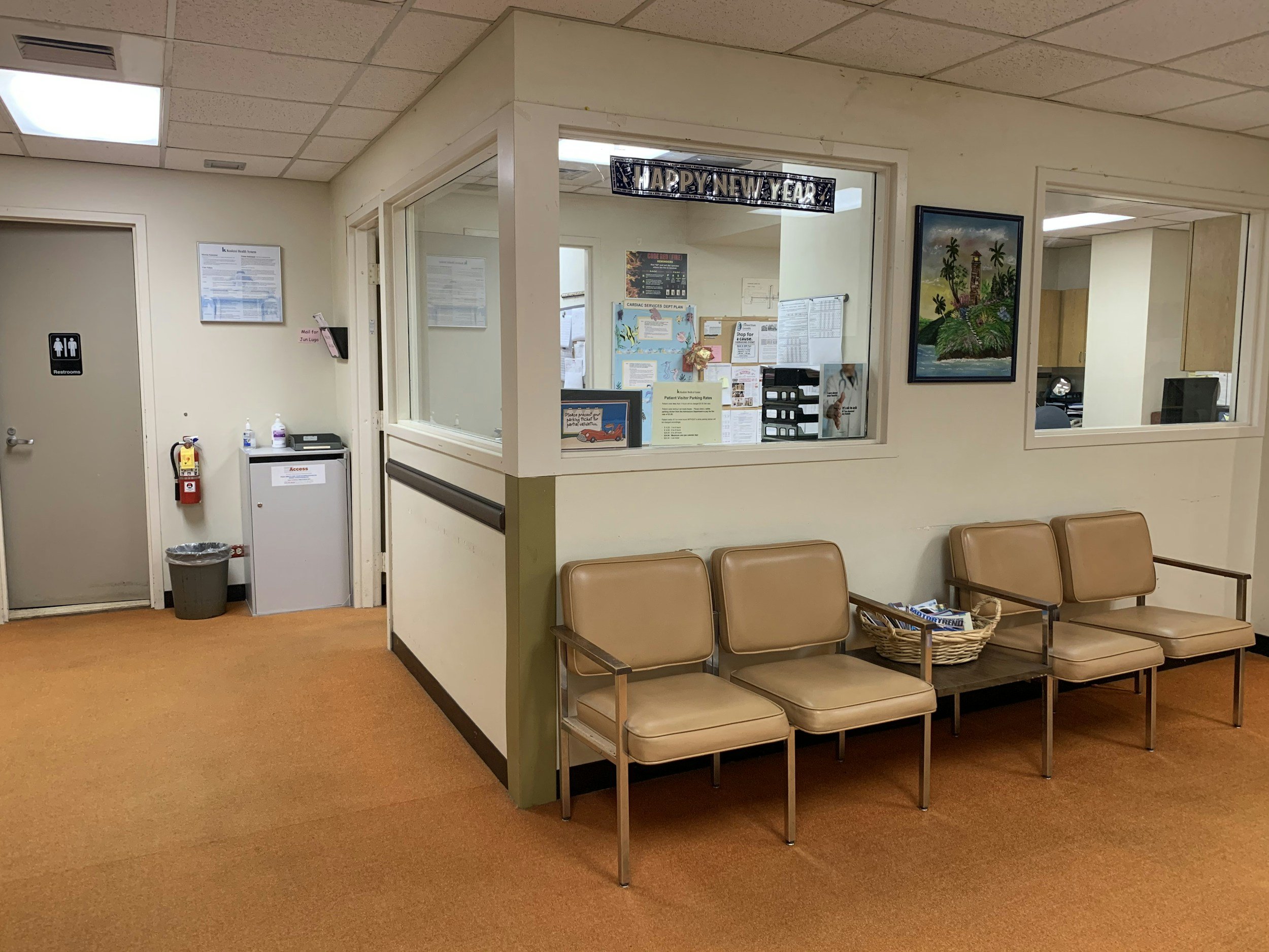 Interior view of a waiting area in a medical facility, with beige chairs arranged in rows, a basket of magazines, a wall with a painting of an island with palm trees, and a reception window behind which informational posters and signs are displayed. A 'Happy New Year' banner hangs above the window.