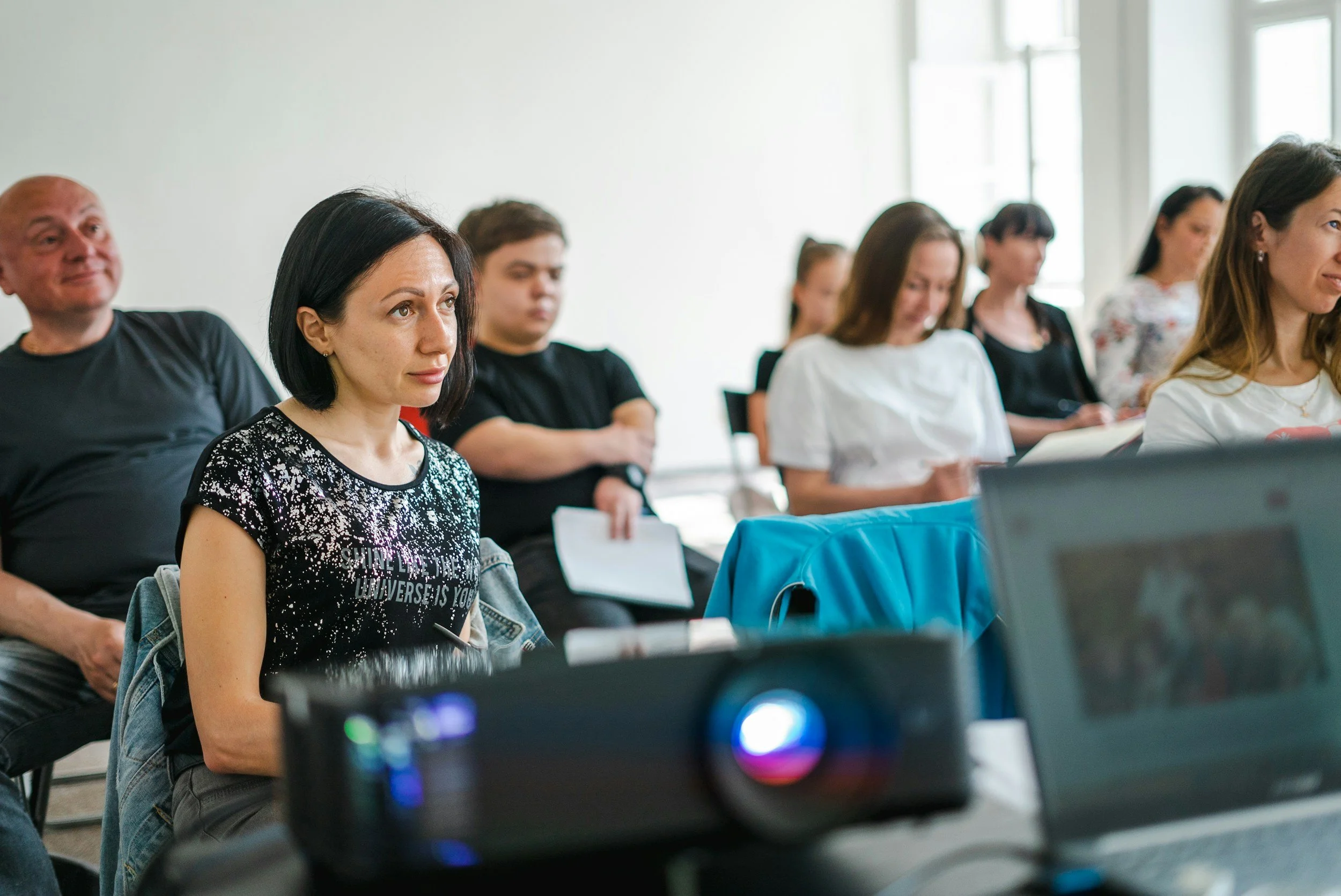 A group of diverse adults seated in a classroom or seminar, attentively listening, some taking notes, with natural light coming in through large windows.