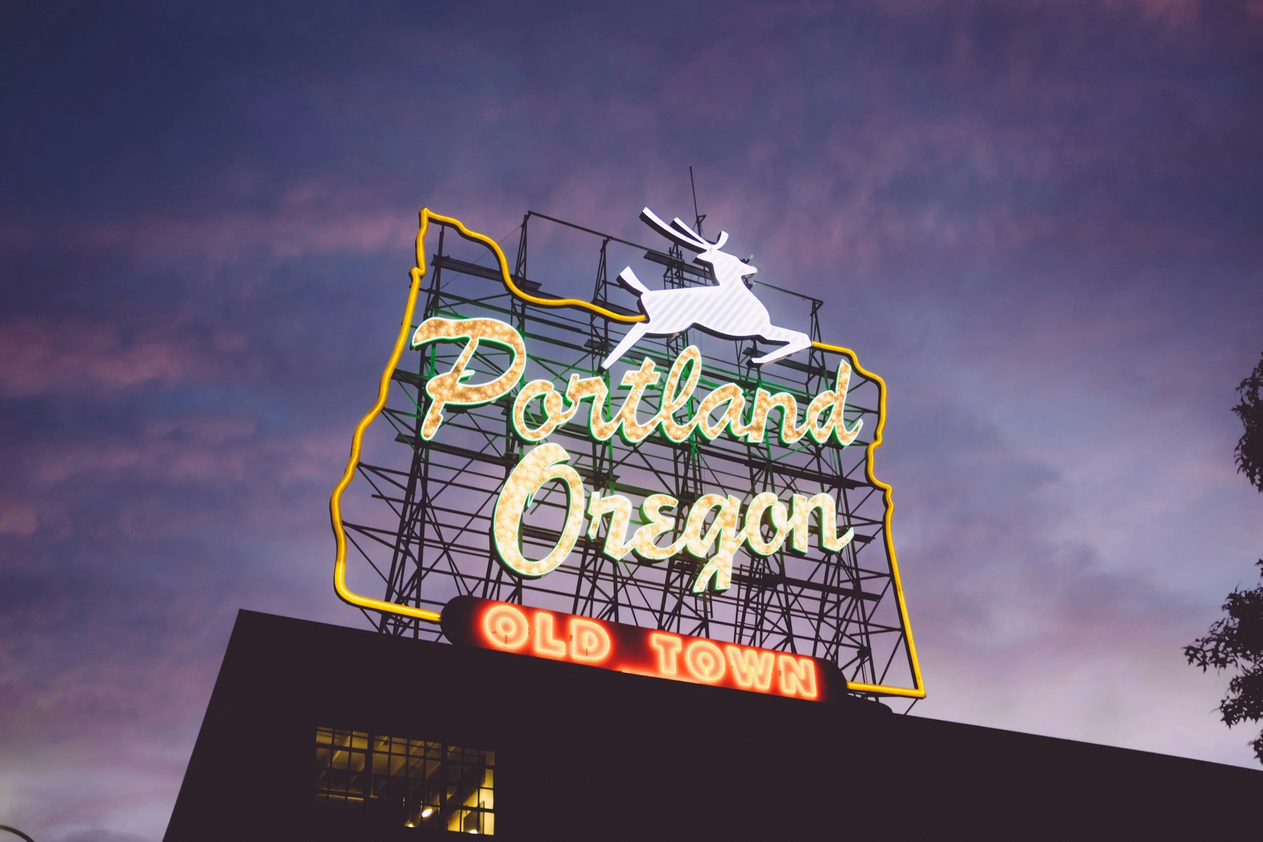 Neon sign reading 'Portland Oregon Old Town' with a white elk or stag on top, set against a darkening sky.