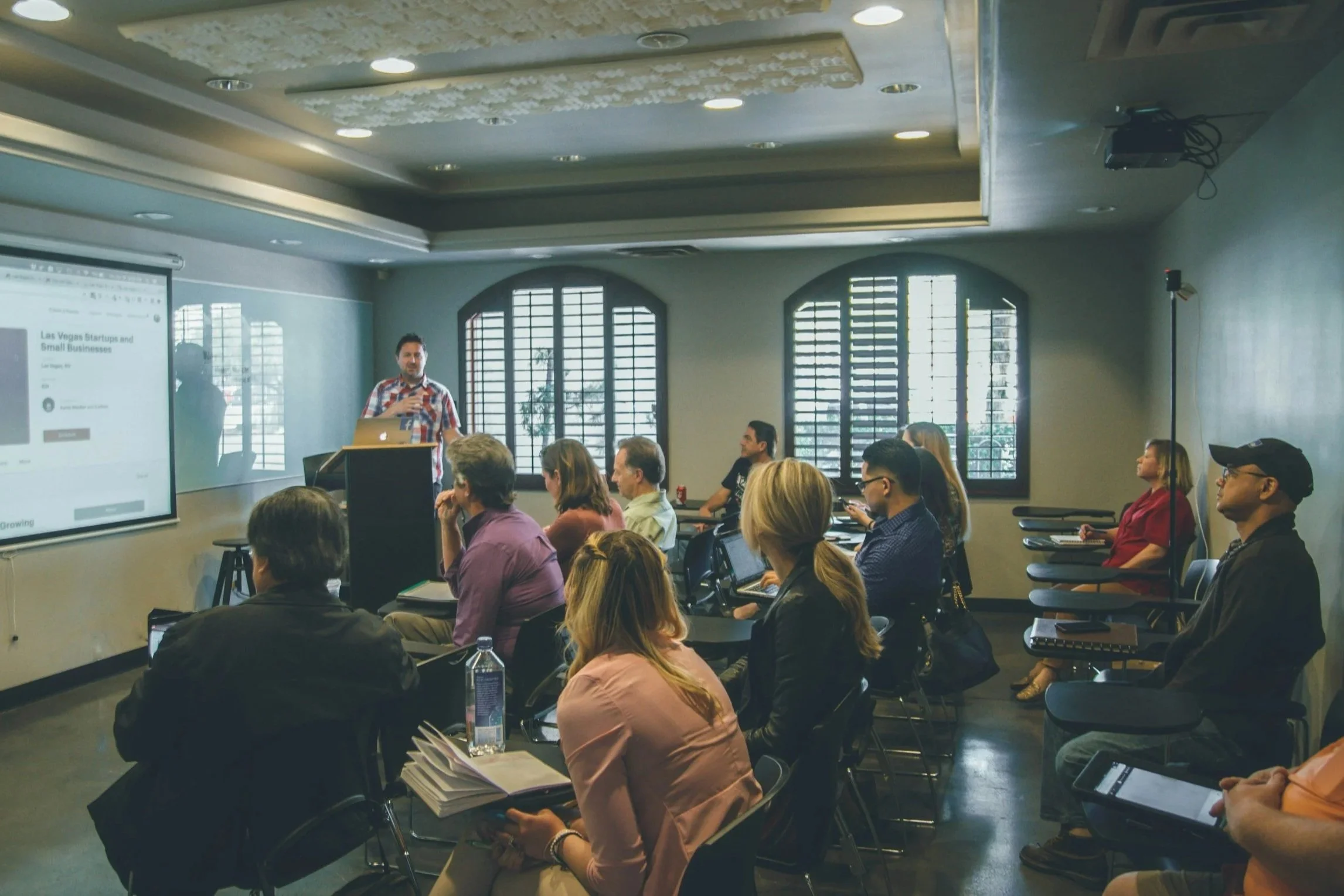 A man giving a presentation to an audience seated in a conference room with large windows and a ceiling mounted projector.
