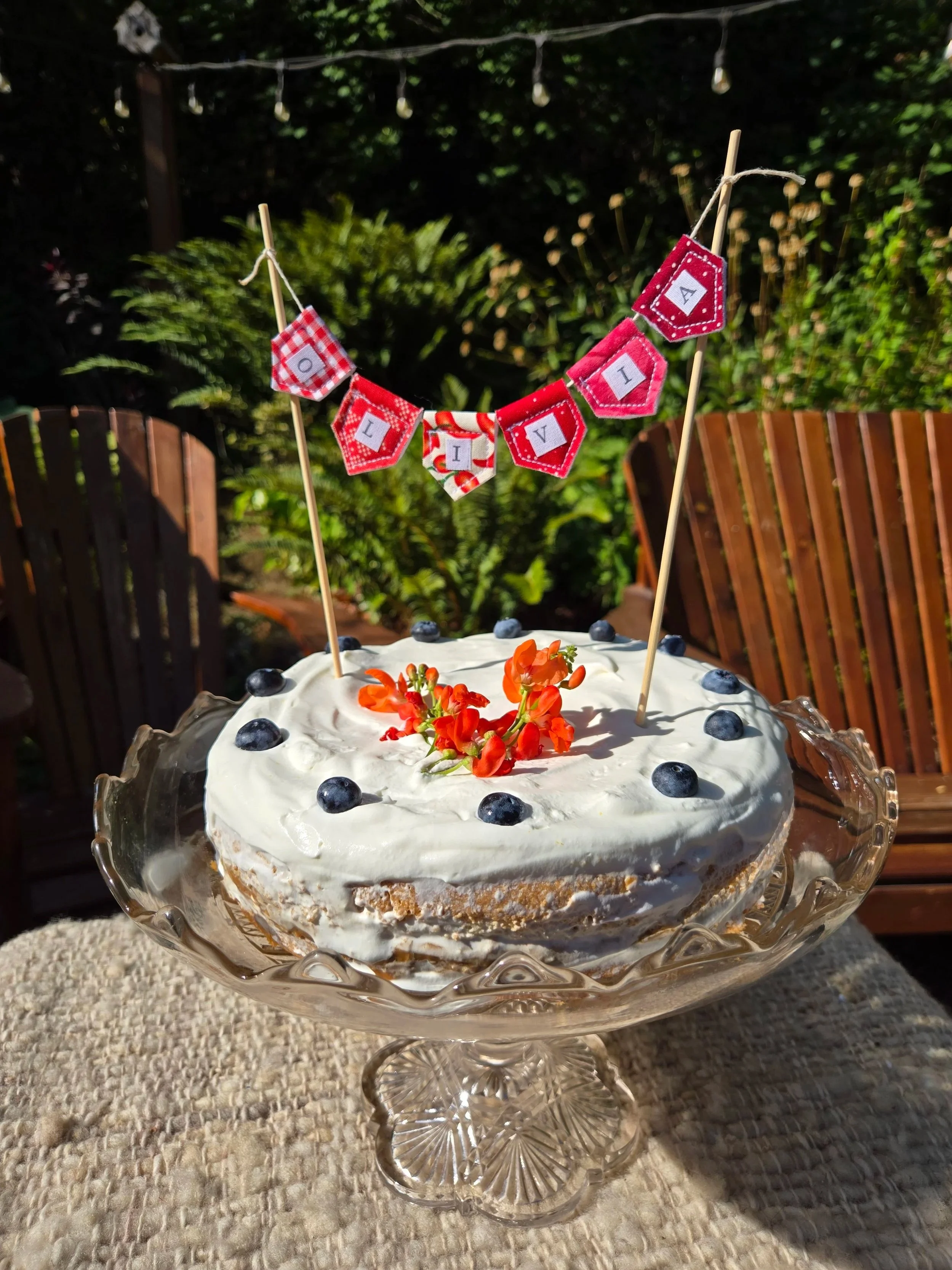 Mini fabric cake topper banner by Birdcraft Studio, made from red reclaimed textiles and styled on a celebration cake decorated with with blueberries.