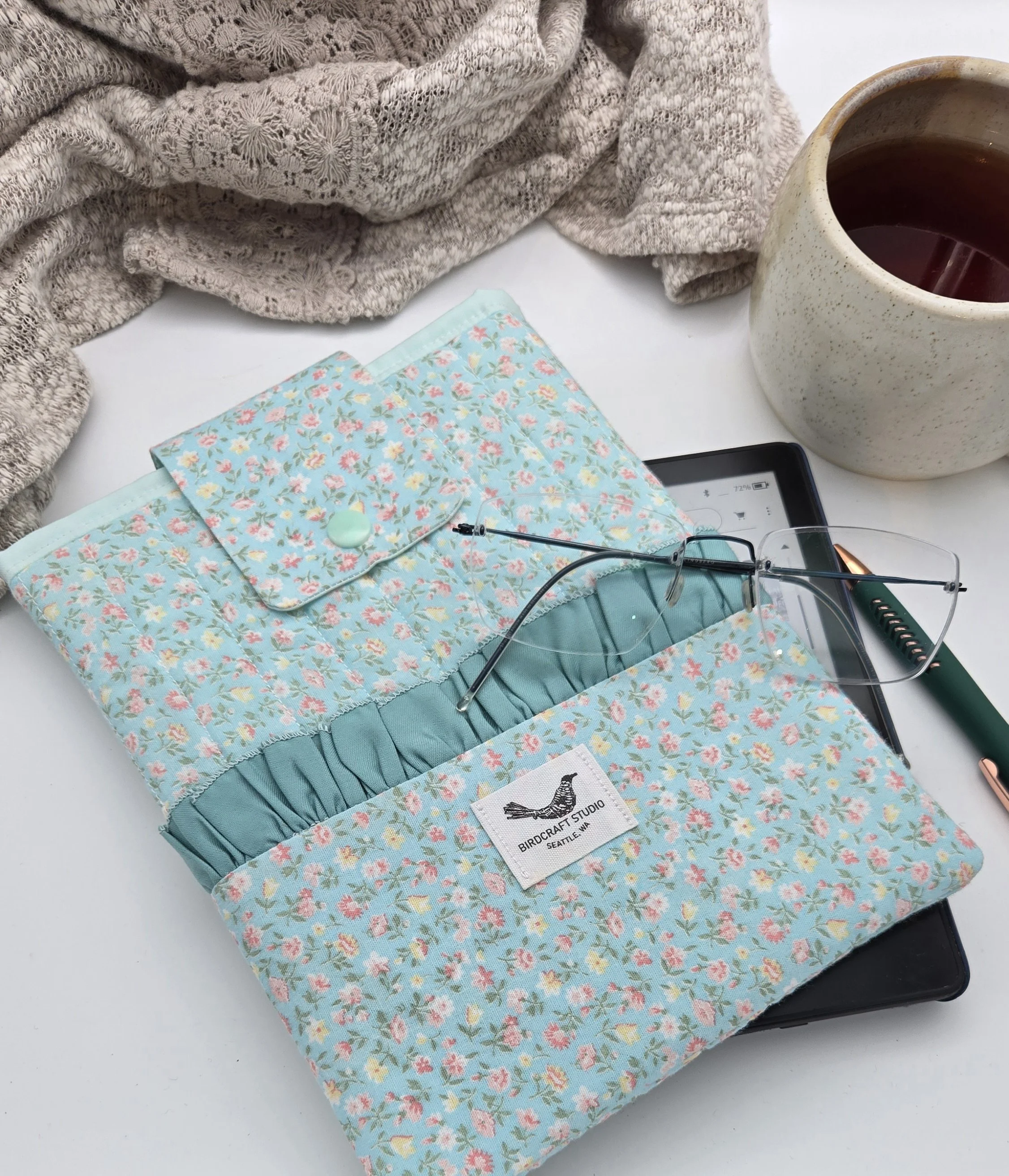Teal ruffled Kindle sleeve with pocket made from upcycled textile, styled with reading glasses, and an e-reader.