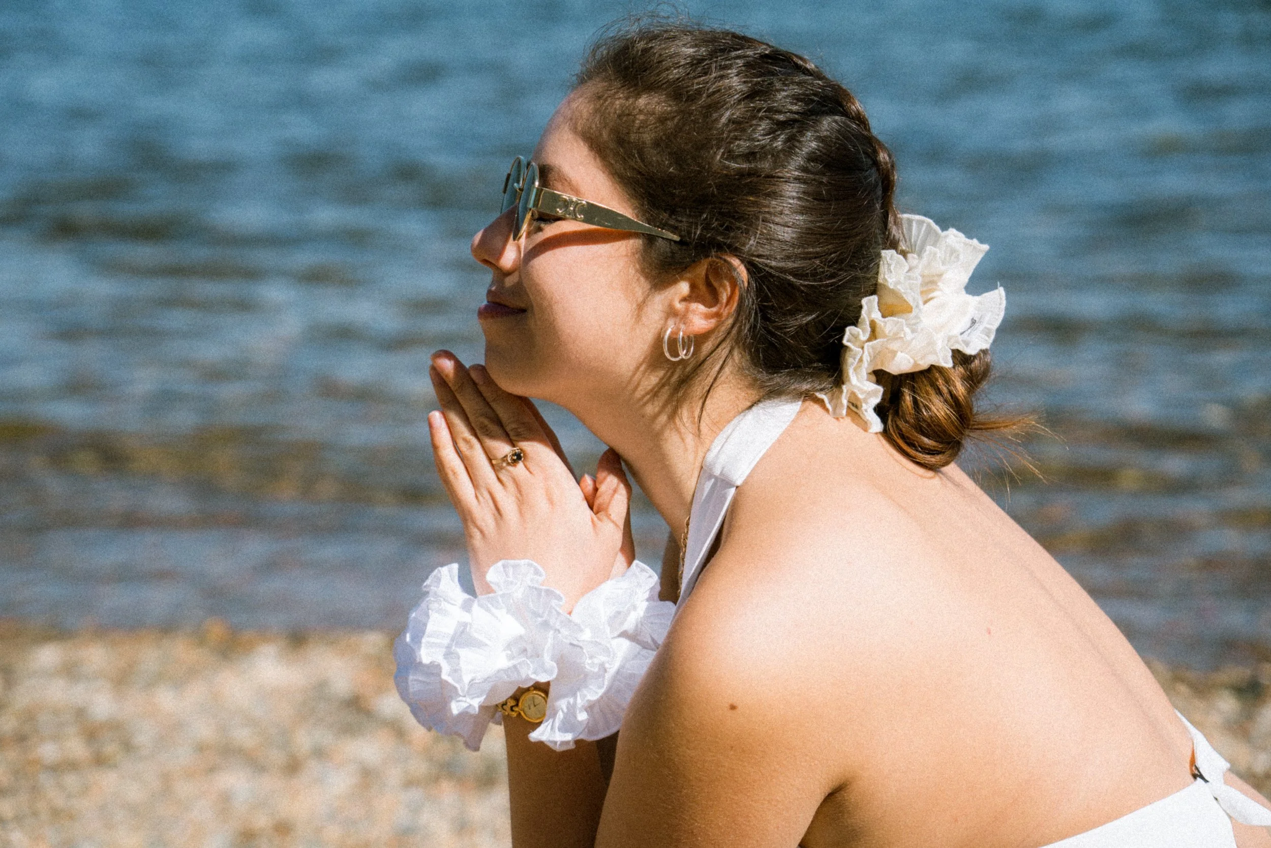 A woman on the beach wears white Birdcraft Studio ruffled scrunchies in her hair and on her wrist, styled with a white dress.