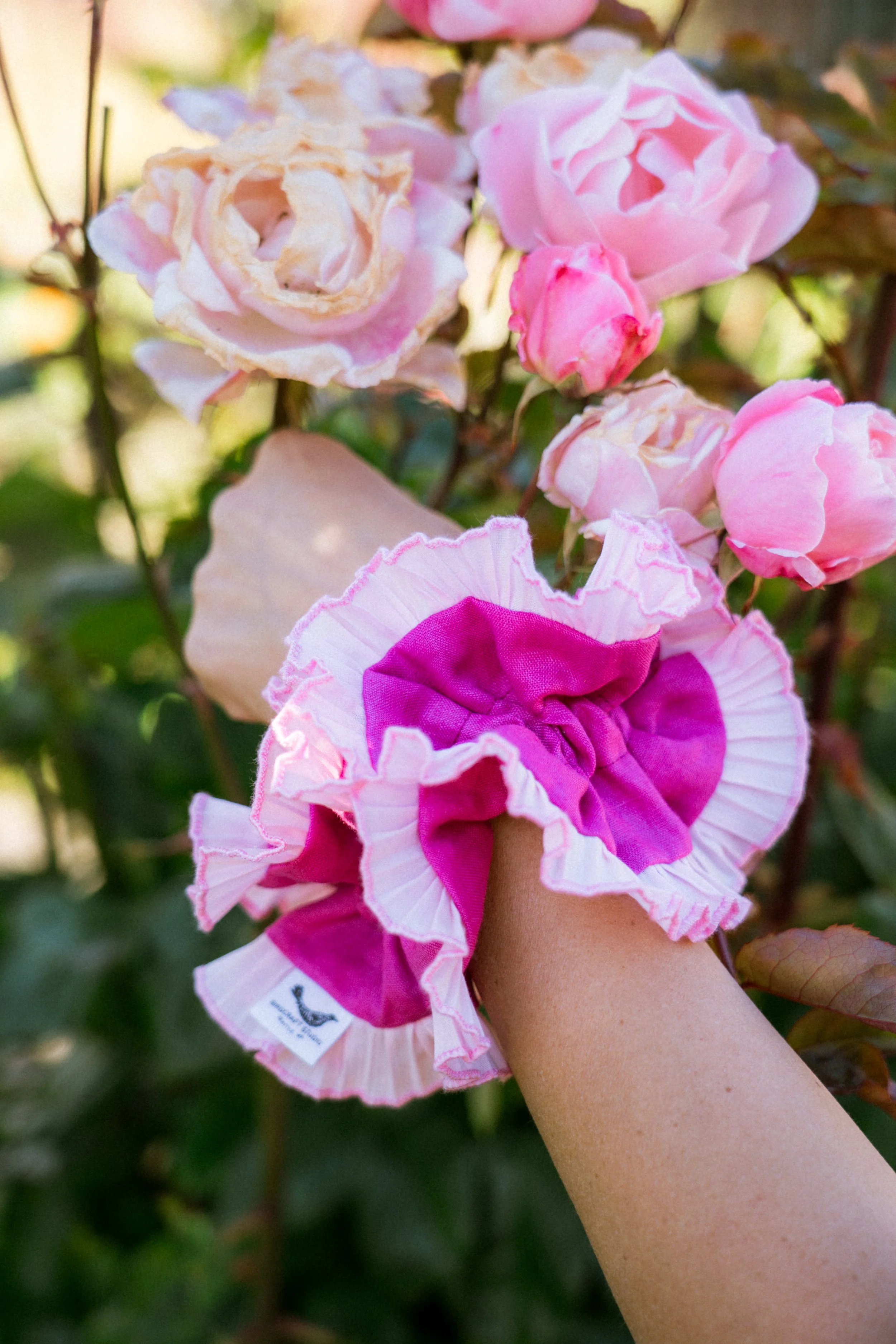 Pink reclaimed-fabric ruffled scrunchie by Birdcraft Studio styled on a wrist beside fresh pink roses in a garden.