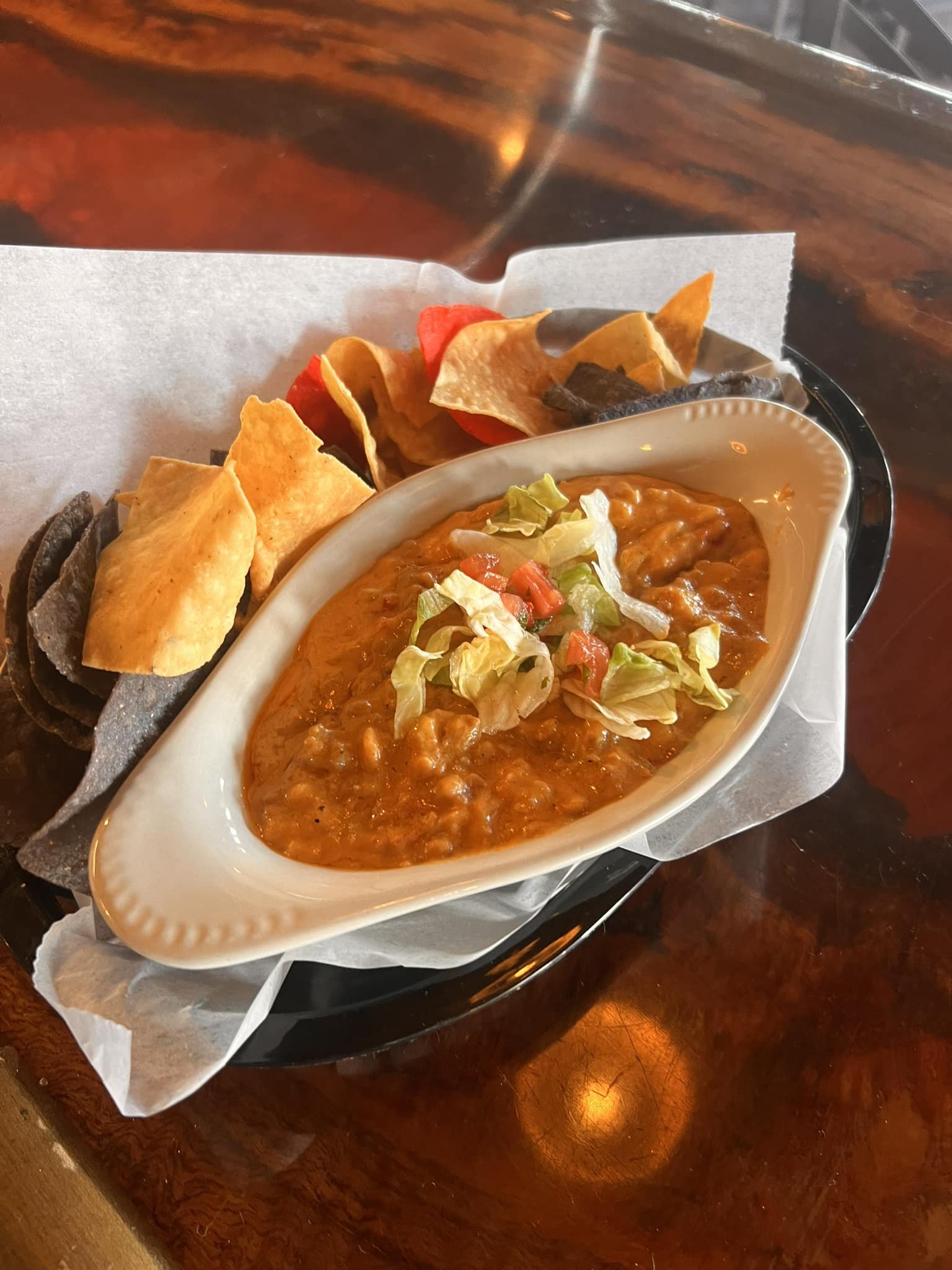 A serving of chili with shredded lettuce on top, alongside assorted tortilla chips in a paper-lined basket, on a black tray.
