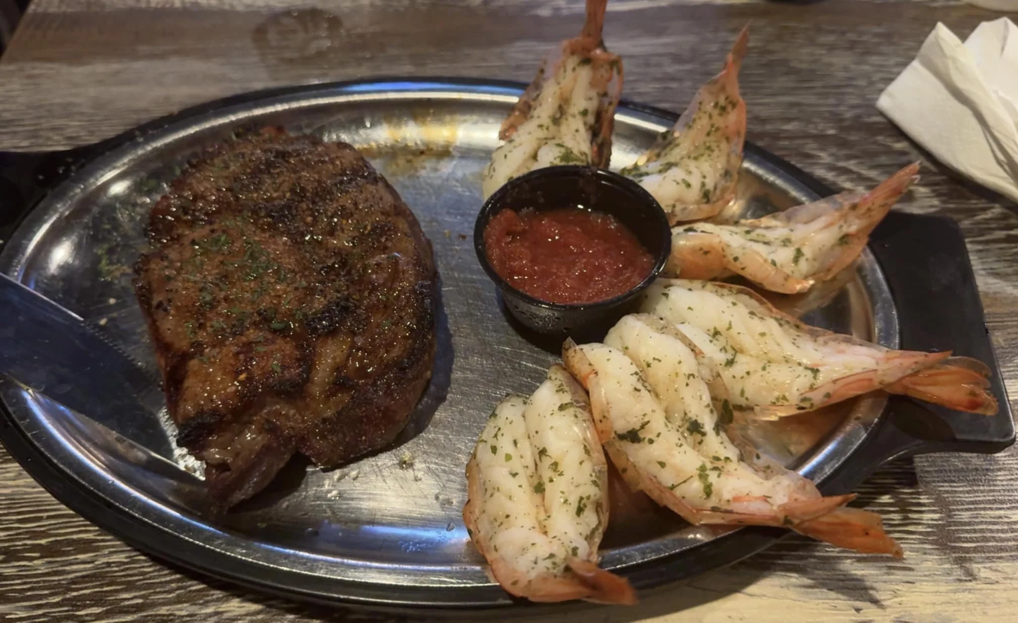 A metal serving tray with a piece of grilled steak, a small cup of red sauce, and several grilled shrimp seasoned with herbs and spices.
