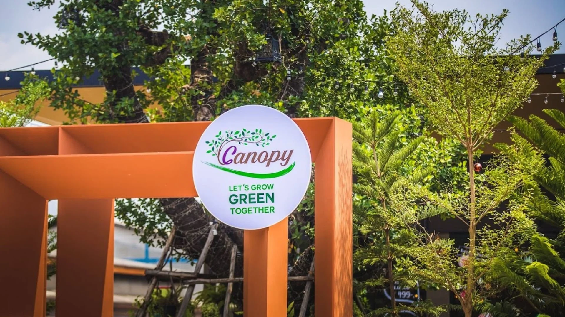 Wooden garden arch with a circular sign that says 'Canopy. Let's Grow Green Together', surrounded by green leafy trees and string lights in the background.
