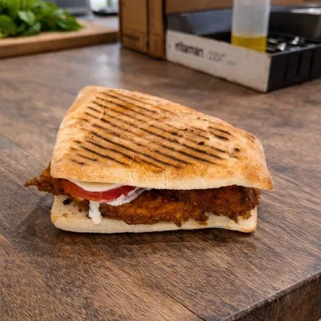 A grilled panini sandwich with bread toasted with grill marks, containing fried chicken, tomato slices, and cheese on a wooden table.