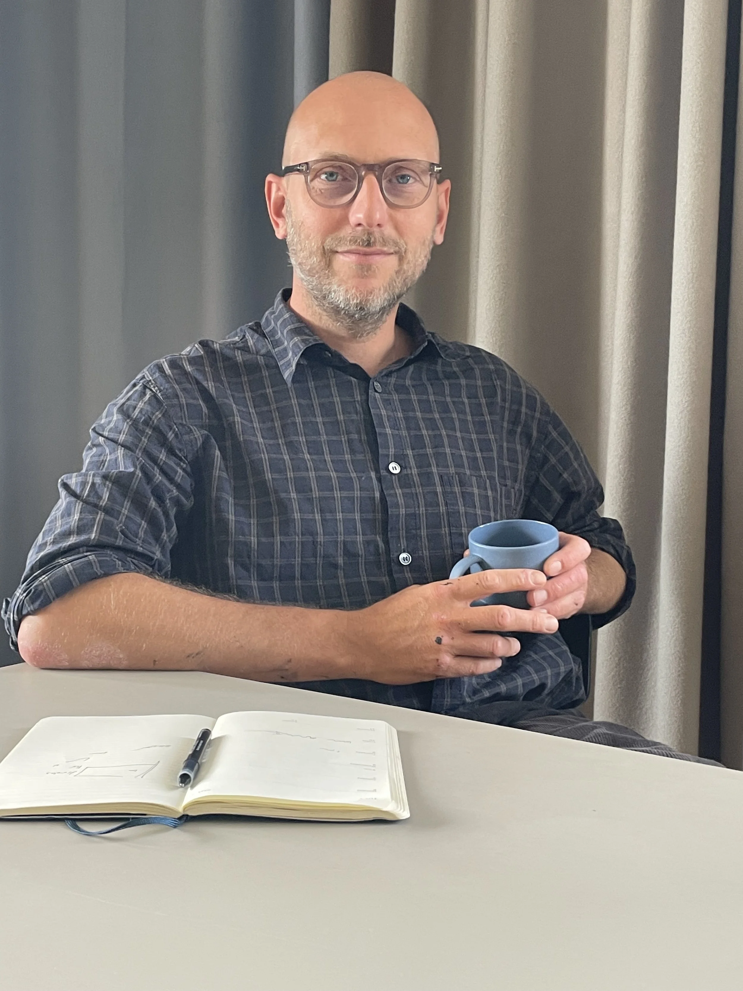 Lasse Bøgeskov Andersson som står bag Pulpo Kommunikation, holder en blå kop, med en åben notesbog og sort pen foran ham, baggrund af gardiner.