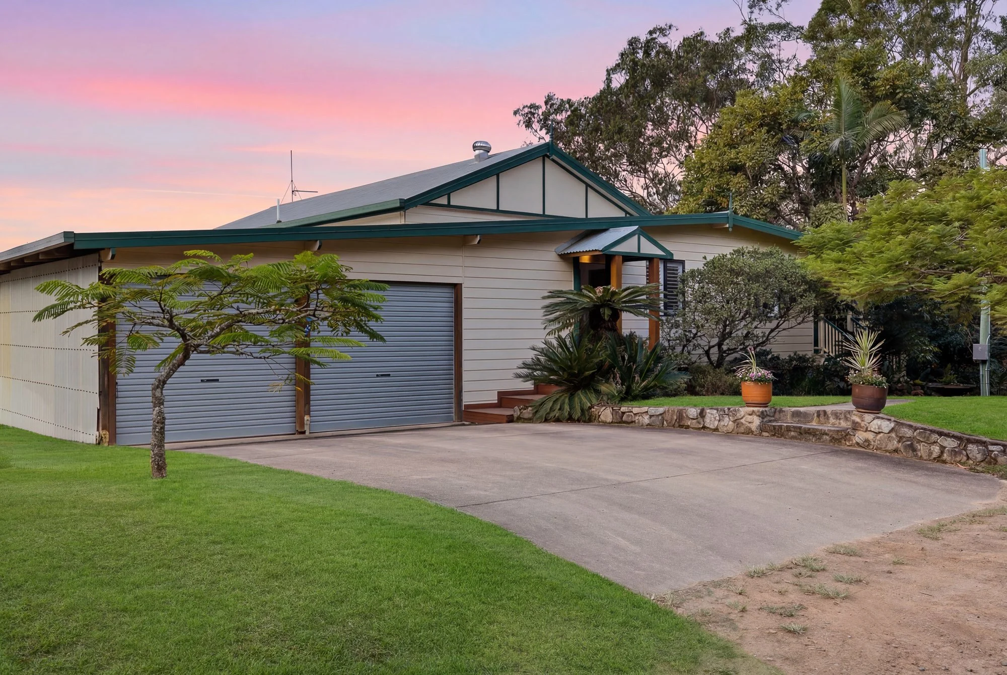 A residential house with a beige exterior, green trim, and a metal roof, surrounded by trees and plants, with a concrete driveway and a sunset sky in the background.