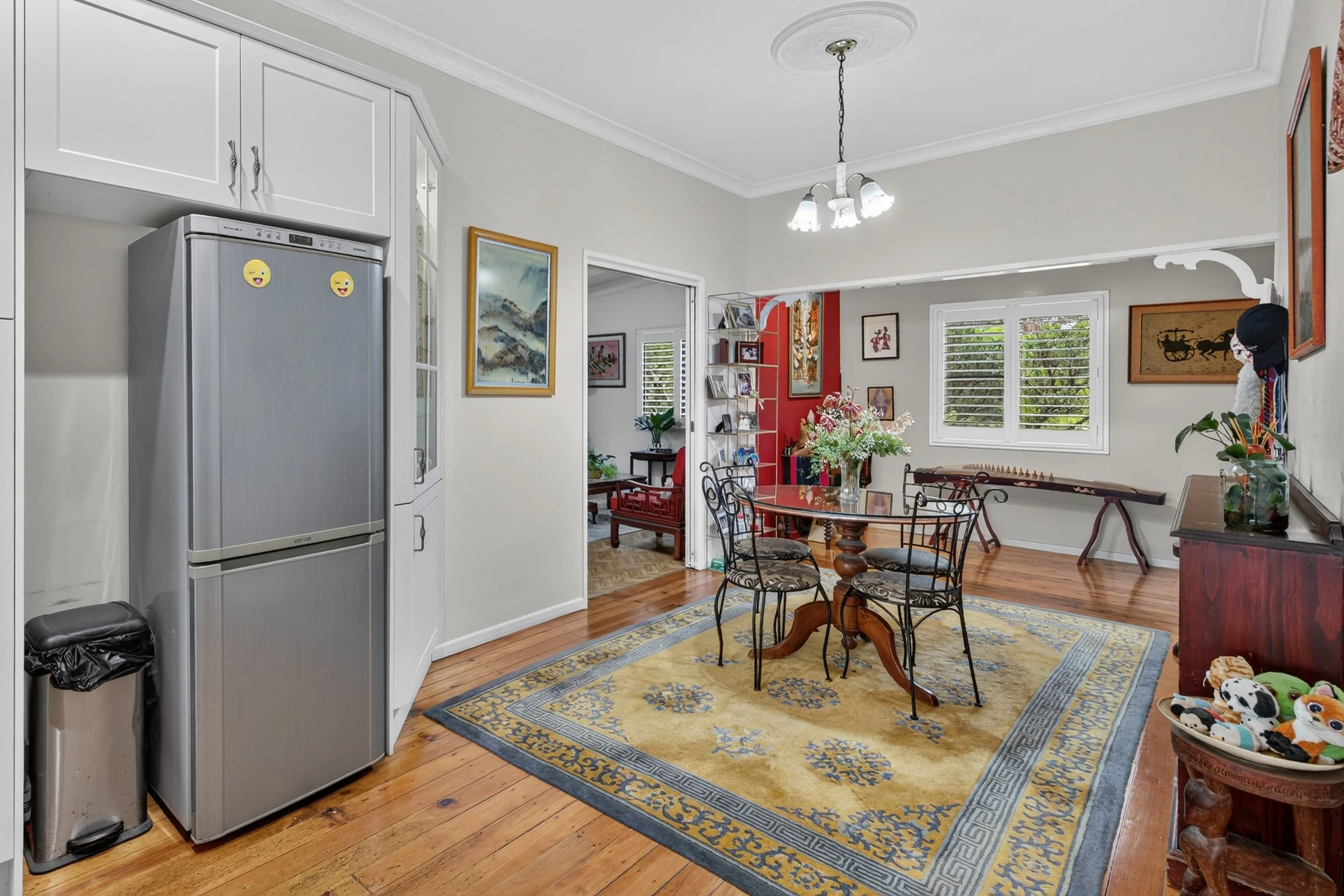 A dining room with wooden floors, a yellow patterned rug, a round wooden table with black metal chairs, and a sideboard. There are windows with white shutters, various framed artwork on the walls, and a floral centerpiece on the table. The space open