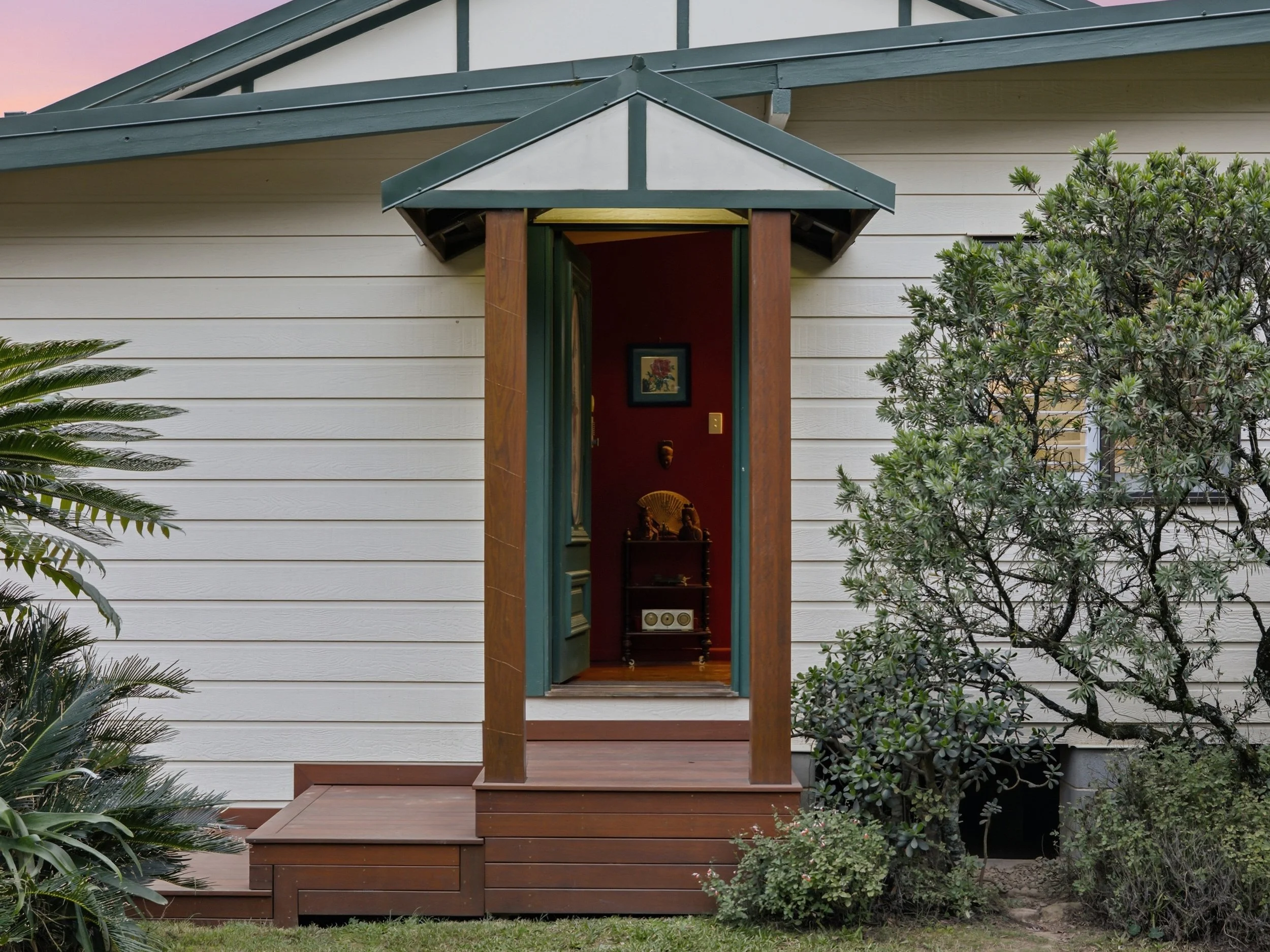 Exterior view of a house with white horizontal siding, green roofing accents, and an entrance with a small porch, surrounded by greenery and trees.