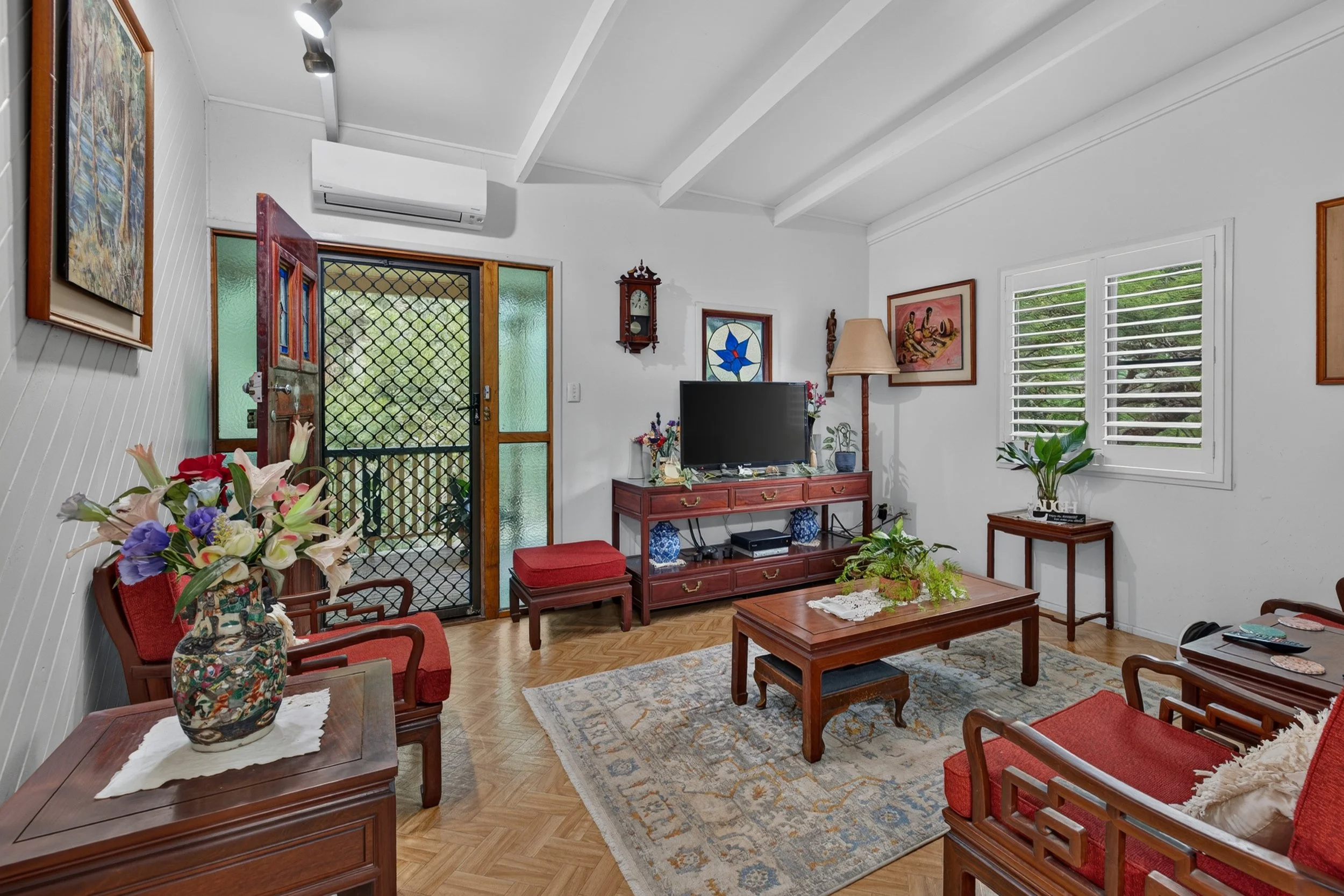 Living room with white walls and ceiling, hardwood floor, red upholstered chairs, wooden coffee table, TV on a wooden stand, floral arrangements, artwork on the walls, and large windows with white shutters.