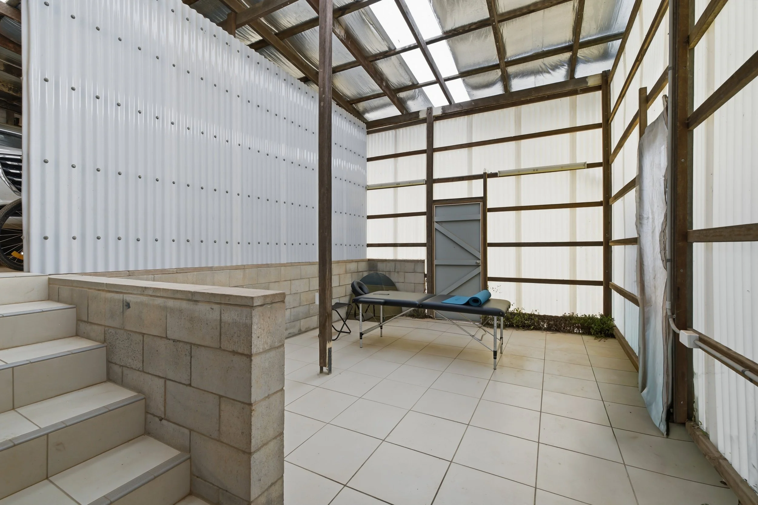 Interior of a partially constructed or renovated building with tiled floor, concrete steps, corrugated metal walls, and a wooden frame ceiling. There is a small folding table with a blue mat and chairs, and a small window on one wall. Some plants are