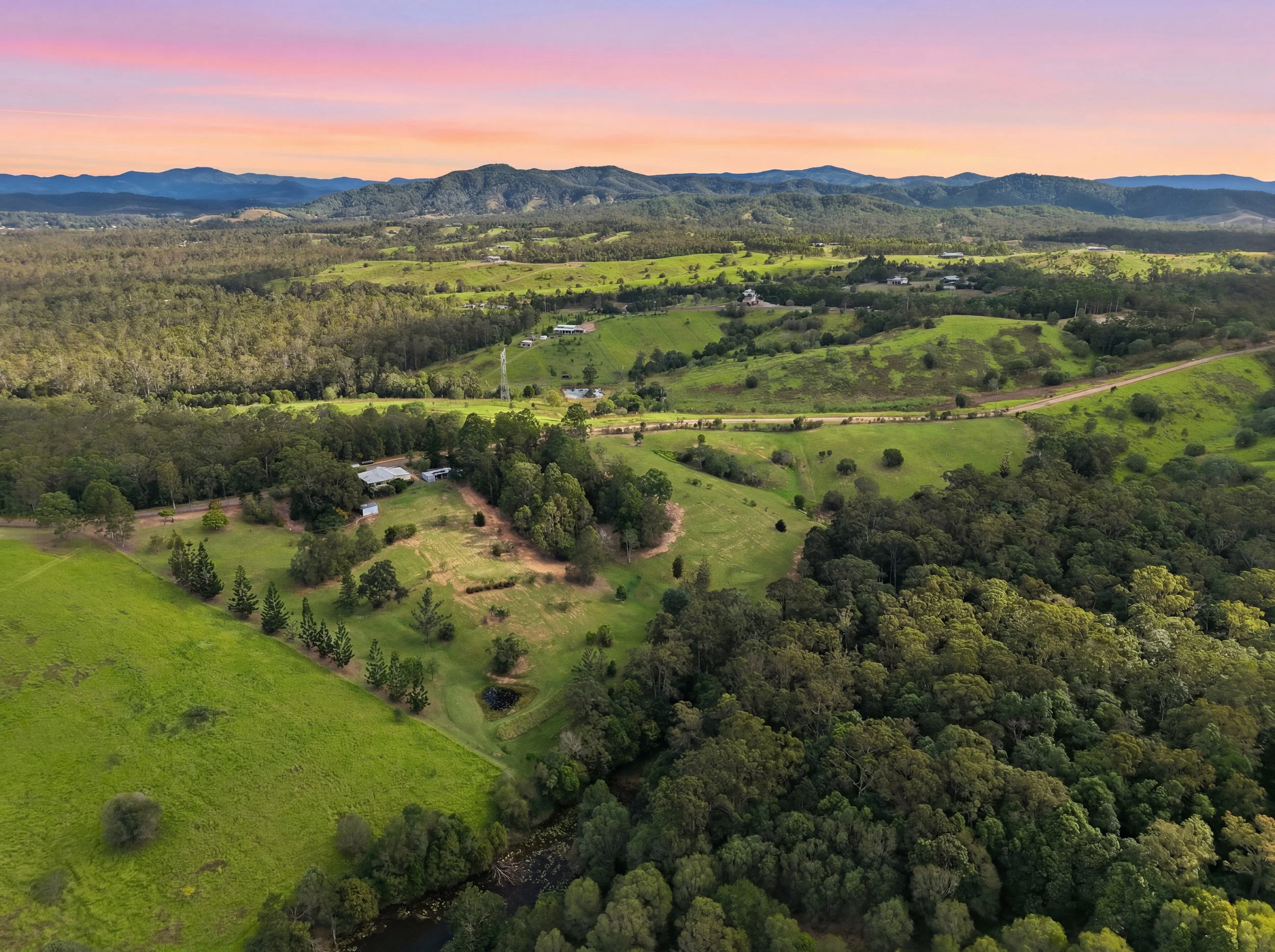A landscape view of rolling green hills, scattered trees, and small houses with a mountain range in the background under a colorful pink and purple sunset sky.