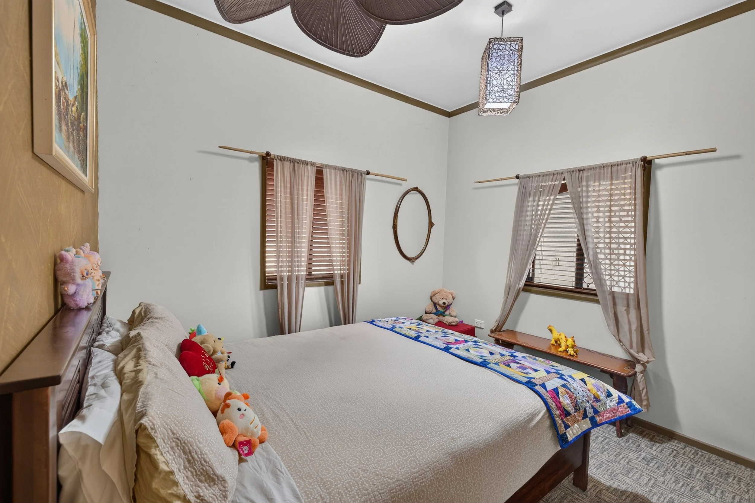 A bedroom with a large bed covered in a textured beige bedspread. Several plush toys are arranged along the head of the bed. There are three windows with wooden blinds and sheer curtains. A quilt with a colorful pattern is draped at the foot of the b