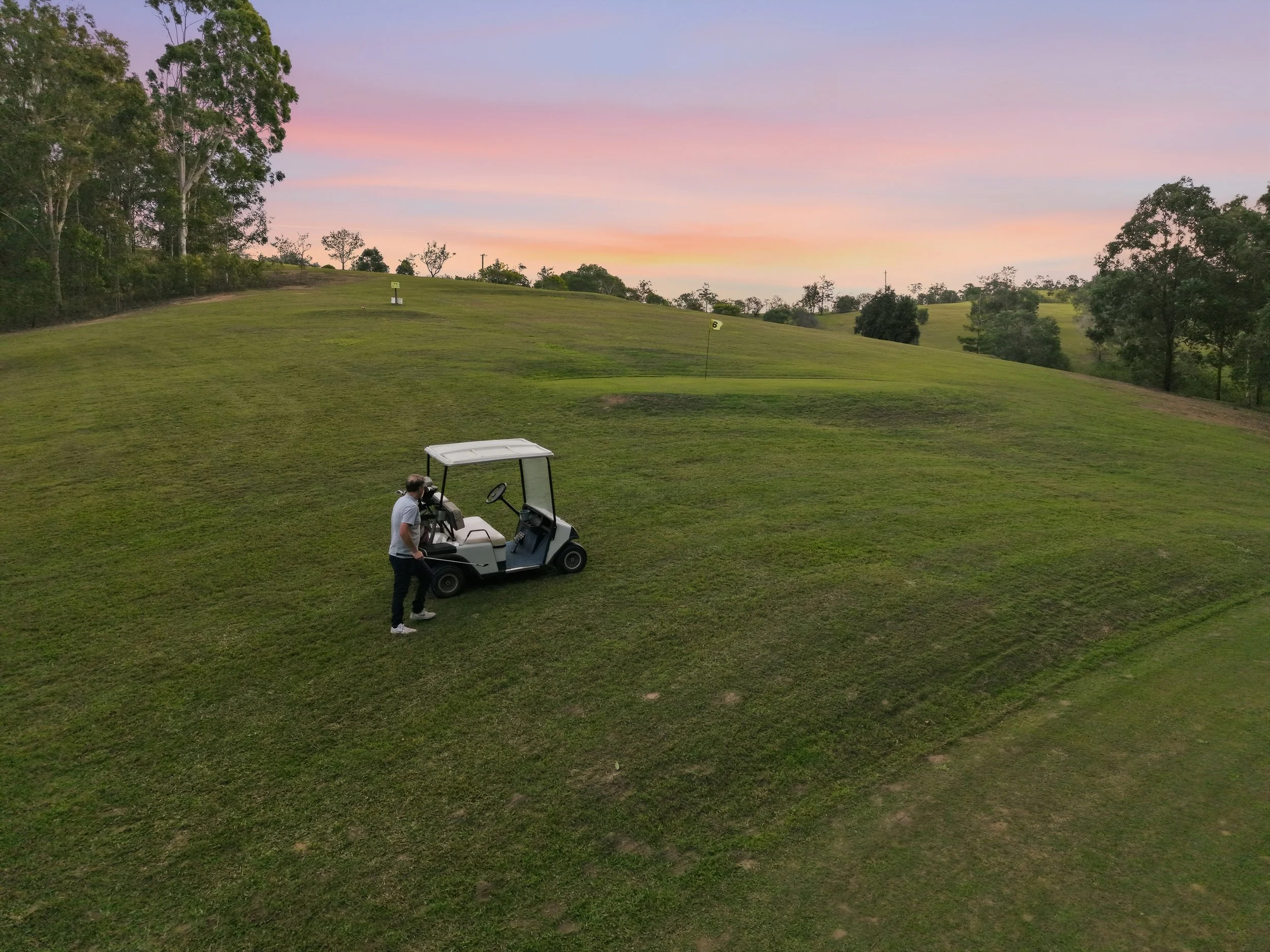 A man in a gray shirt and black pants standing next to a white golf cart on a grassy golf course during sunset, with trees in the background and a pink and purple sky.