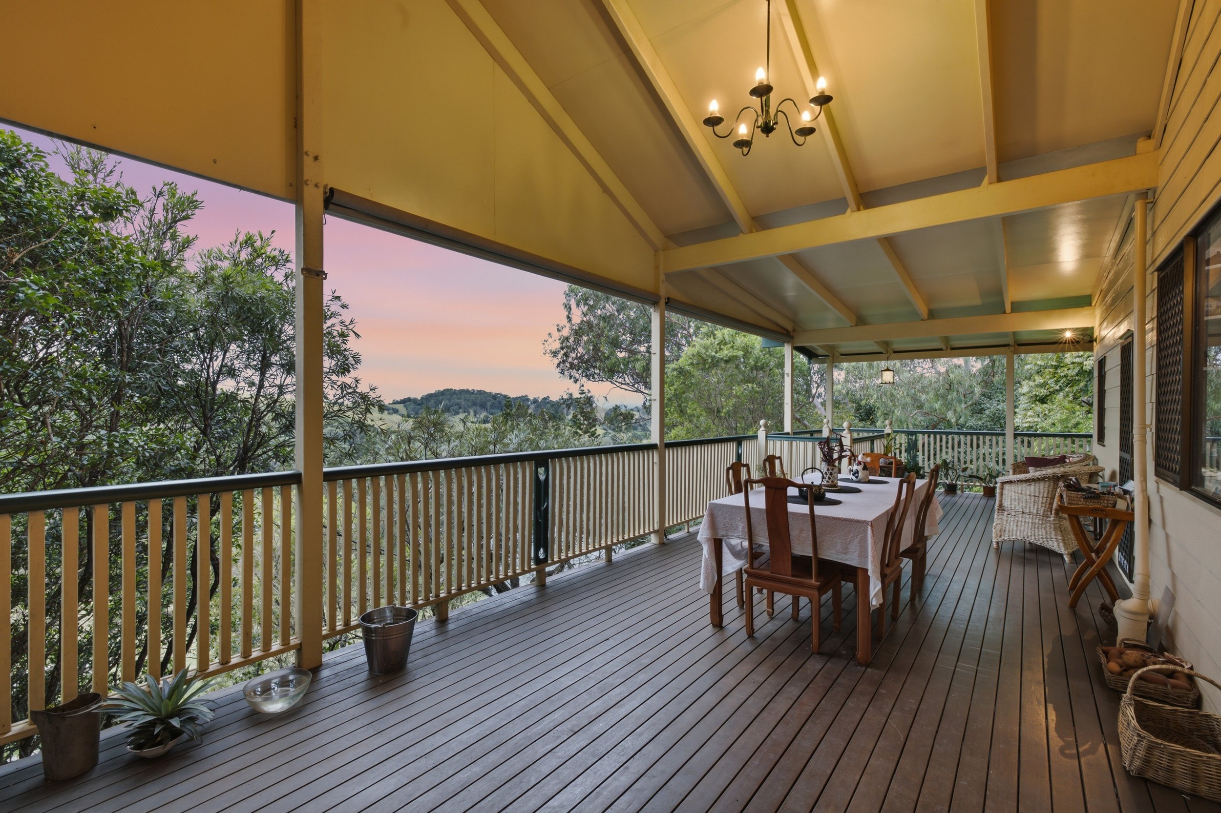 Covered outdoor porch with wooden flooring, a dining table with chairs, wicker seating, and a chandelier, surrounded by trees during sunset.
