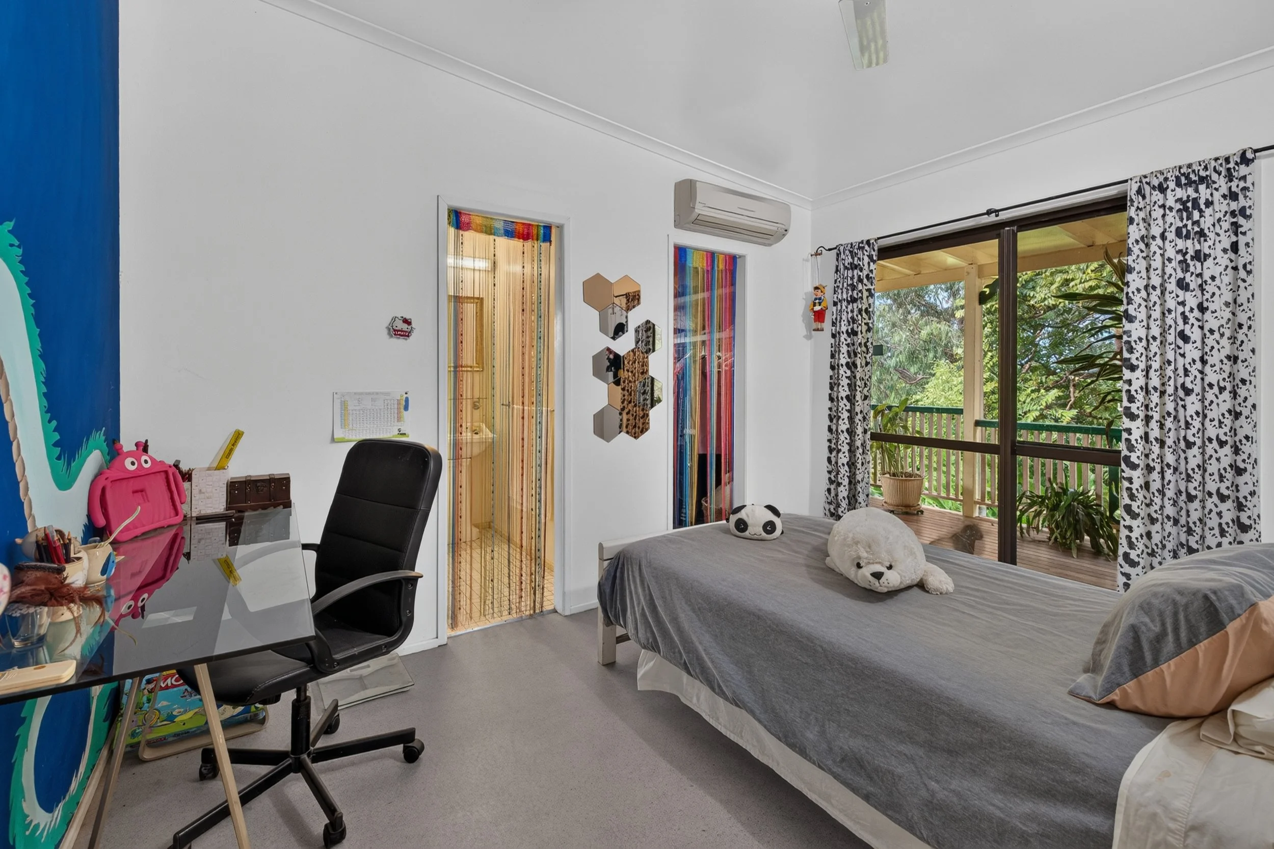 A bedroom with a bed, stuffed animal, and a large window showing a balcony and trees outside. There's a desk with a pink backpack, a black office chair, and wall decorations including colorful beads and mirrored hexagons.