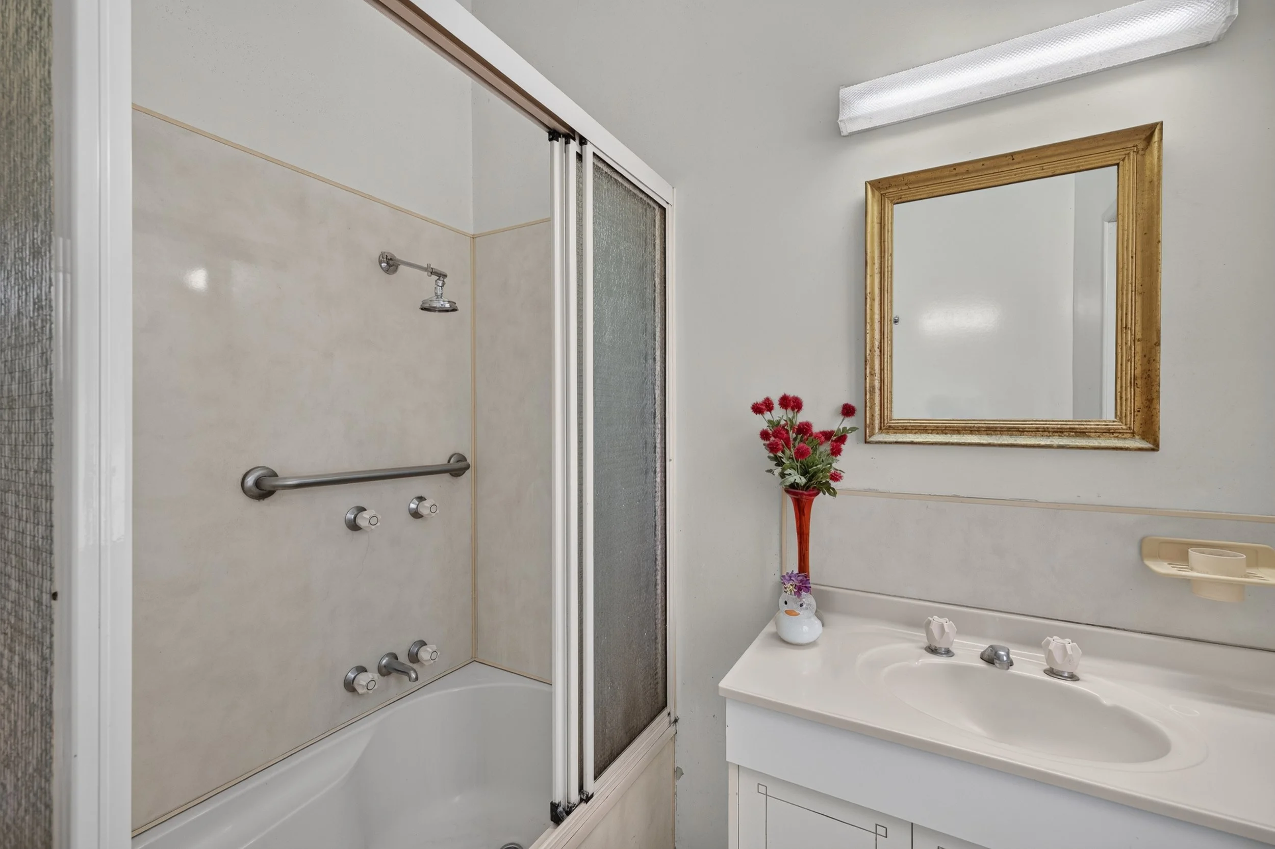 A bathroom with a shower area, including a showerhead and a grab bar, a sink with a mirror, and a vase with pink flowers on the counter.